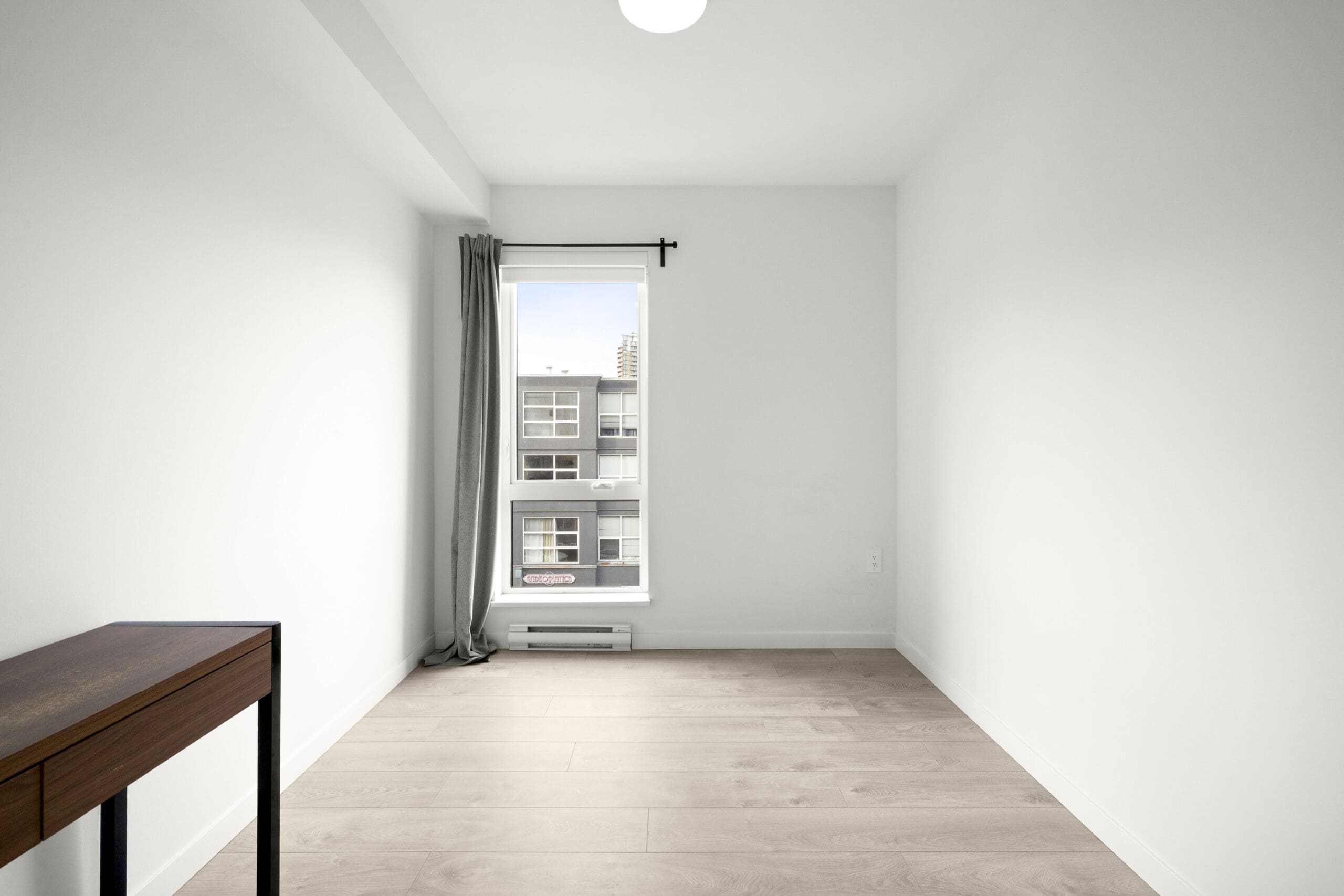 A small, empty room with light-colored flooring, a single window with gray curtains, white walls, and a narrow wooden table on the left side.