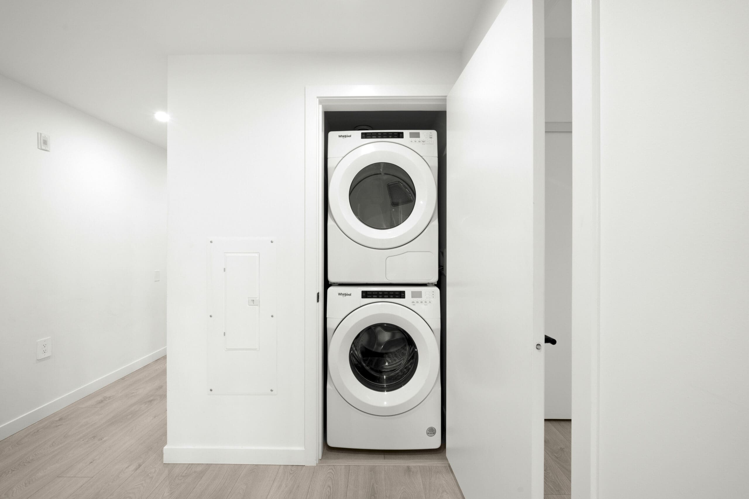 A stacked washer and dryer are installed in a small, white laundry closet with the door partially open in a modern, minimalist room.