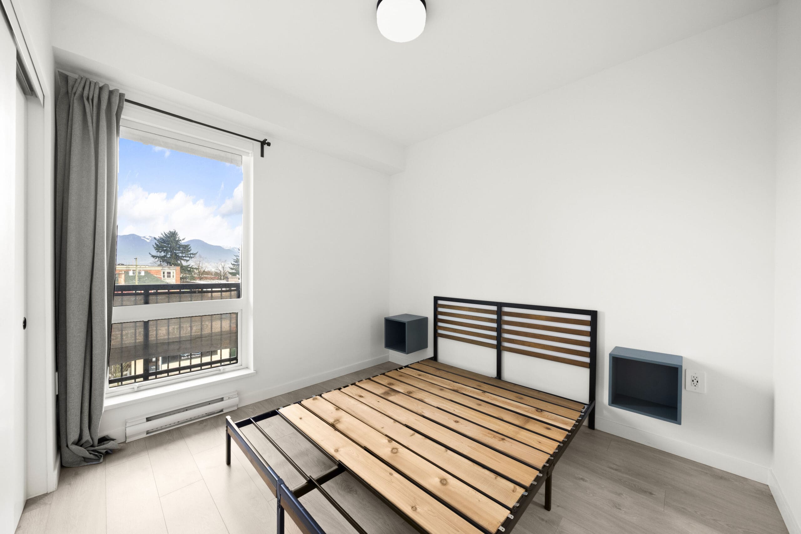 Minimalist bedroom with a wooden bed frame, two wall-mounted nightstands, a window with gray curtains, and white walls.