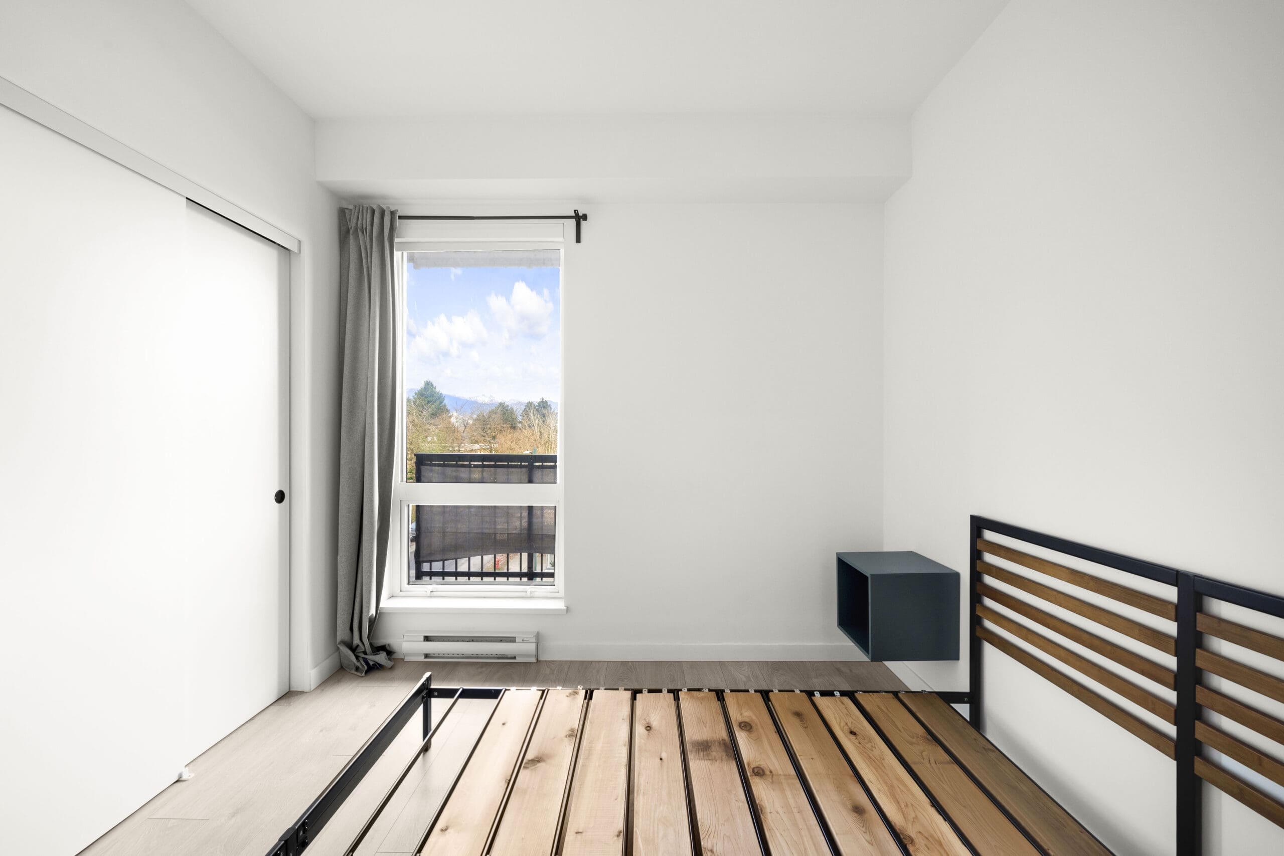 Minimalist bedroom with a window showing a view of trees, an empty bed frame, white walls, gray curtains, and a sliding closet door.