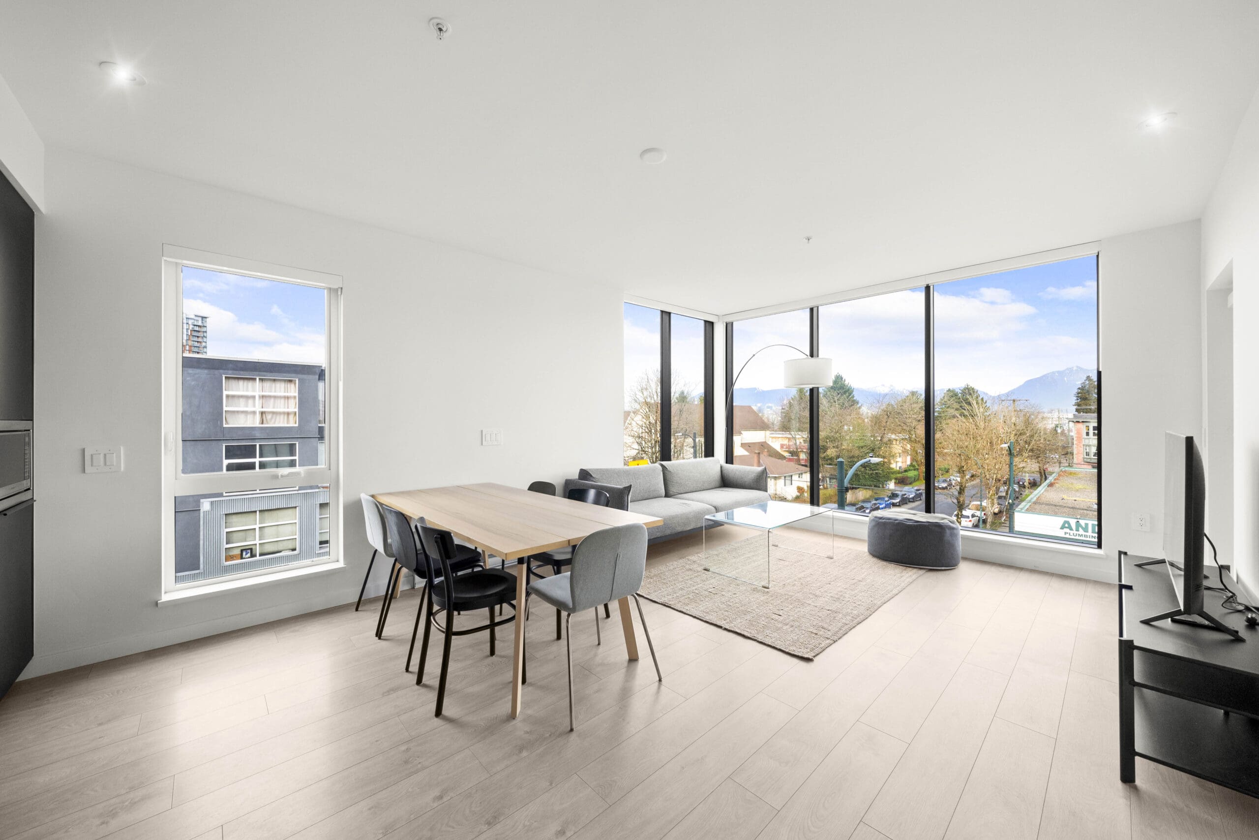 Modern, bright living and dining area with large corner windows, light wood flooring, a dining table with chairs, sectional sofa, TV, and views of city buildings and mountains.