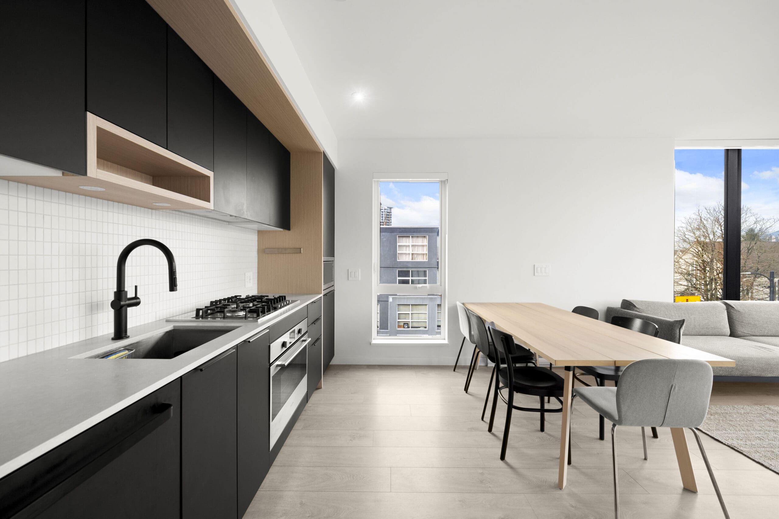Modern kitchen with black cabinets, built-in appliances, a long wooden dining table with chairs, and a living area with a sofa near large windows.