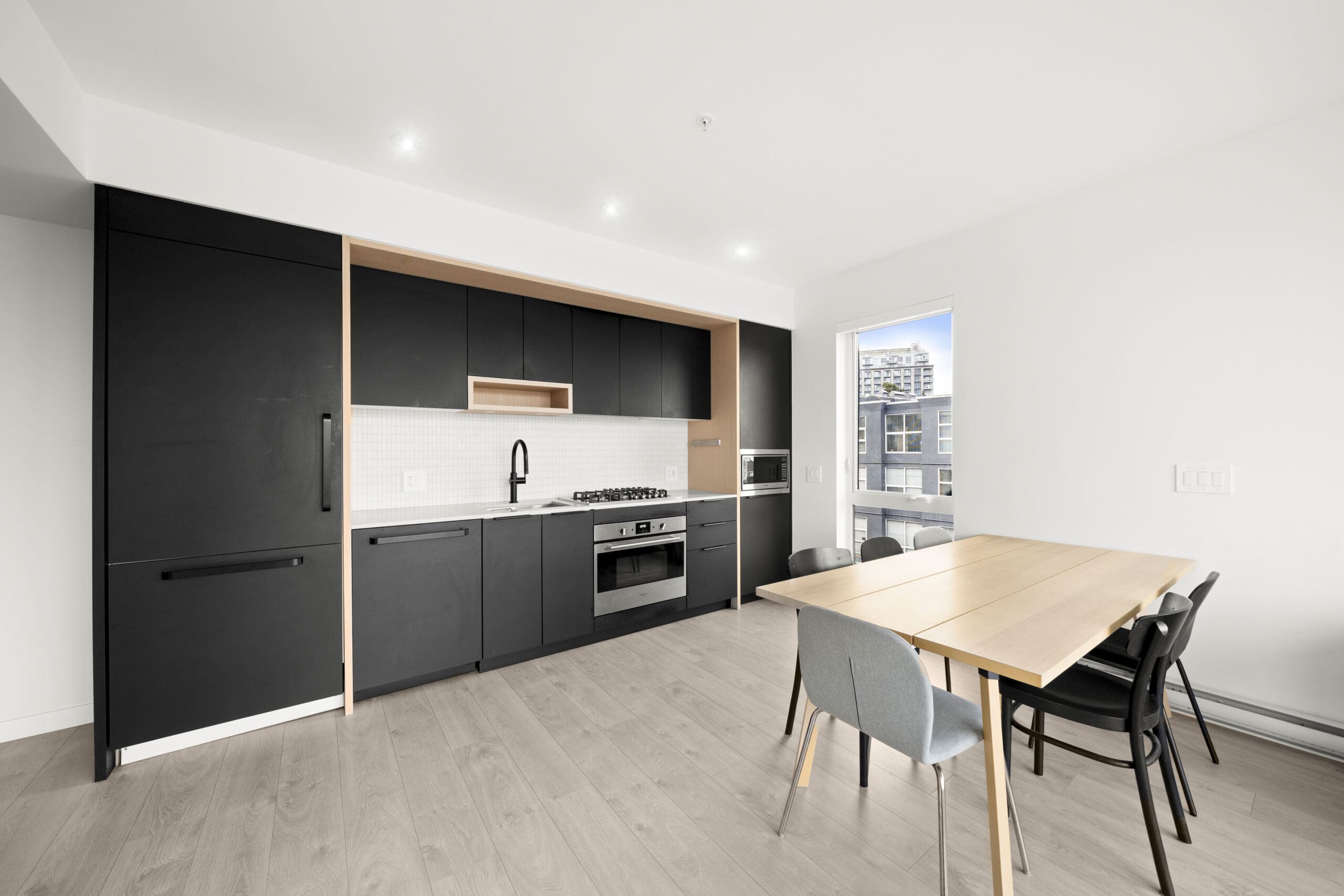 Modern kitchen with black cabinets, built-in appliances, and a light wood dining table with four chairs in a bright room with large window.