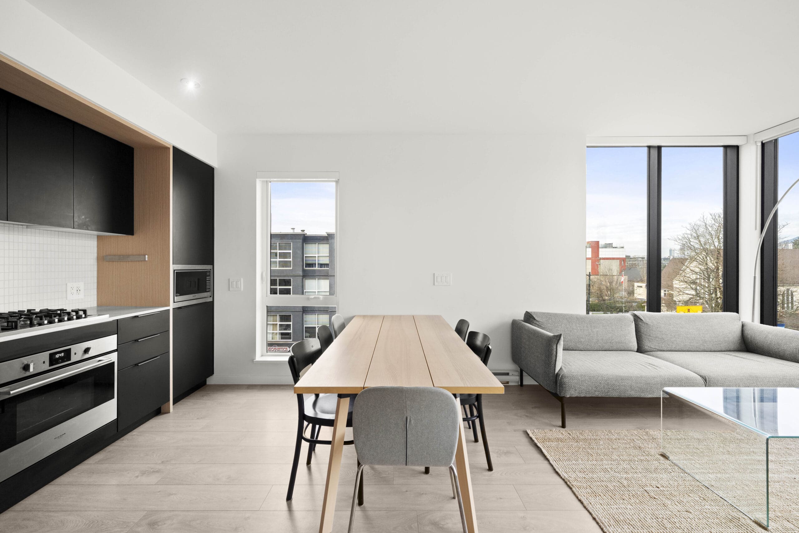 Modern kitchen and dining area with black cabinets, wood dining table with six chairs, gray sofa, large windows, and city views.