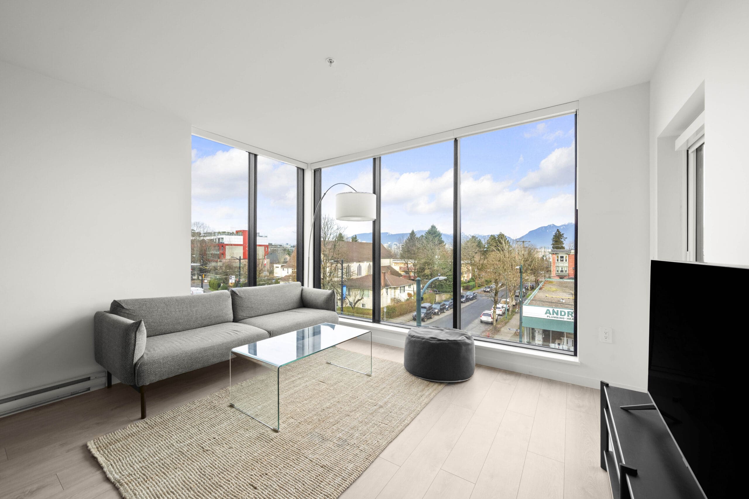 Modern living room with grey sofa, glass coffee table, rug, large floor-to-ceiling windows overlooking a cityscape, and a TV on a stand.