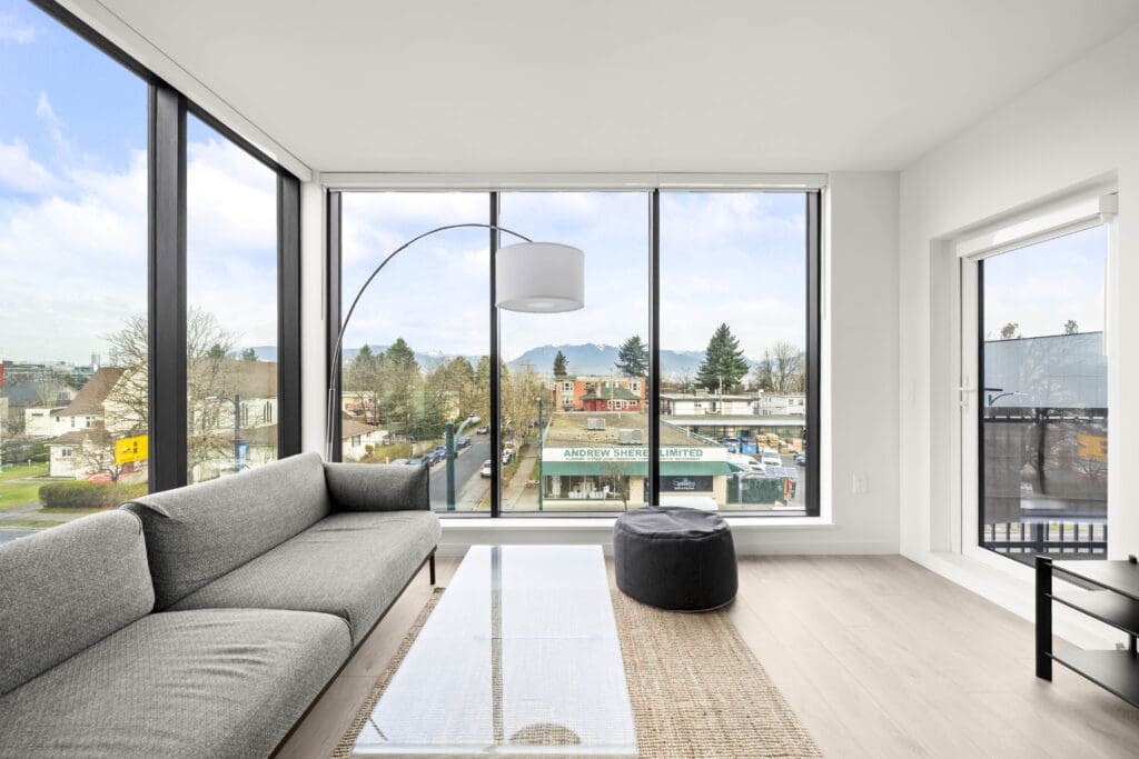 Modern living room with large windows, gray sofa, glass coffee table, and a view of shops and trees outside. Natural light fills the bright, minimalist space.