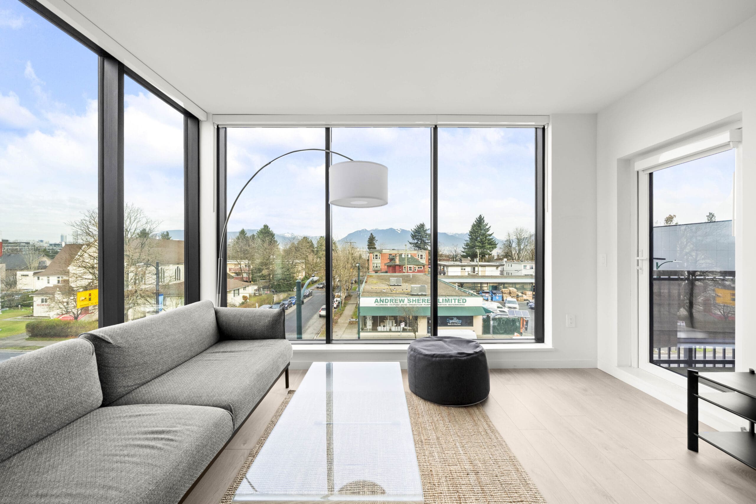 Modern living room with large windows, gray sofa, glass coffee table, and a view of shops and trees outside. Natural light fills the bright, minimalist space.