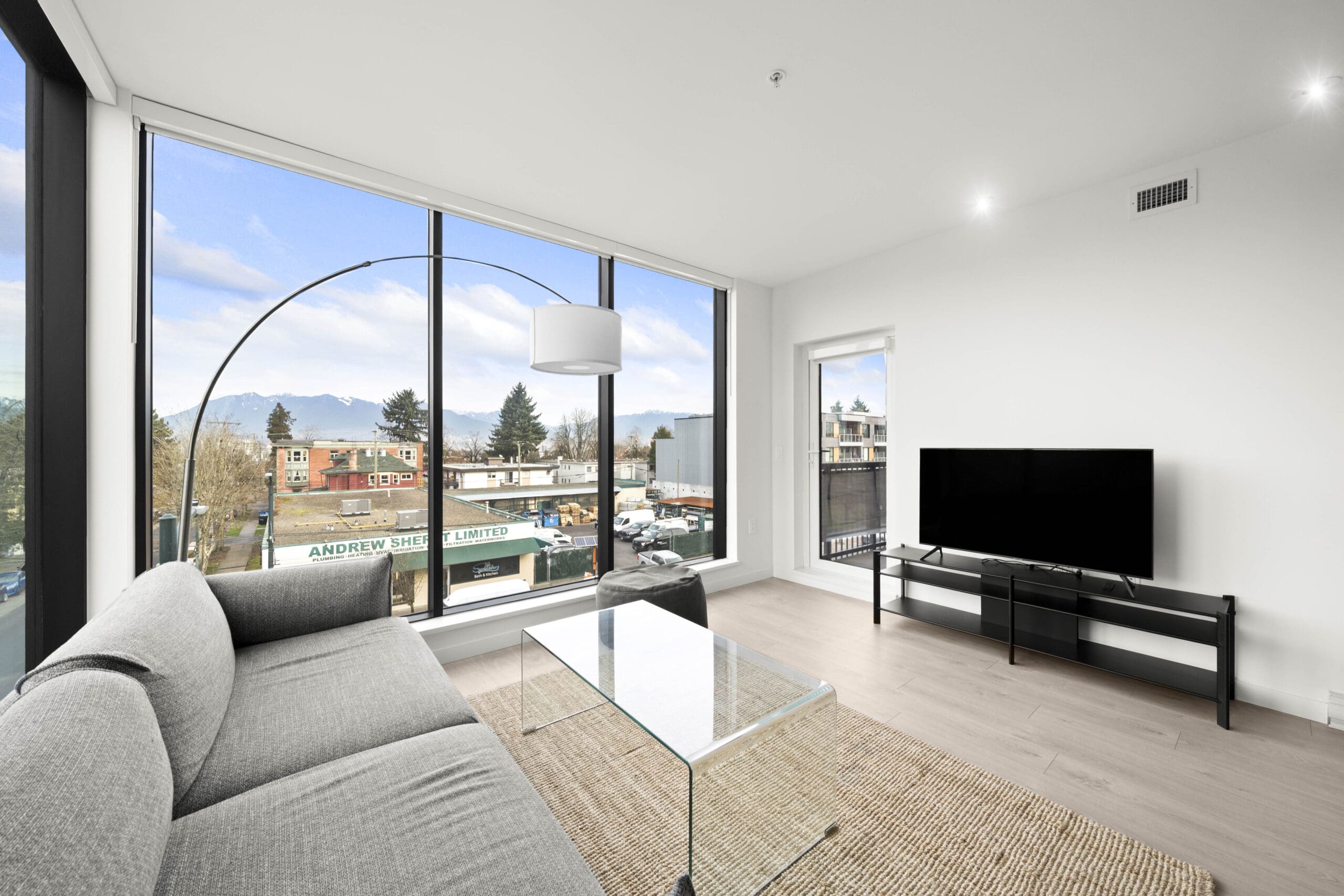 Modern living room with large floor-to-ceiling windows, a gray sofa, glass coffee table, TV on a stand, and cityscape view outside.