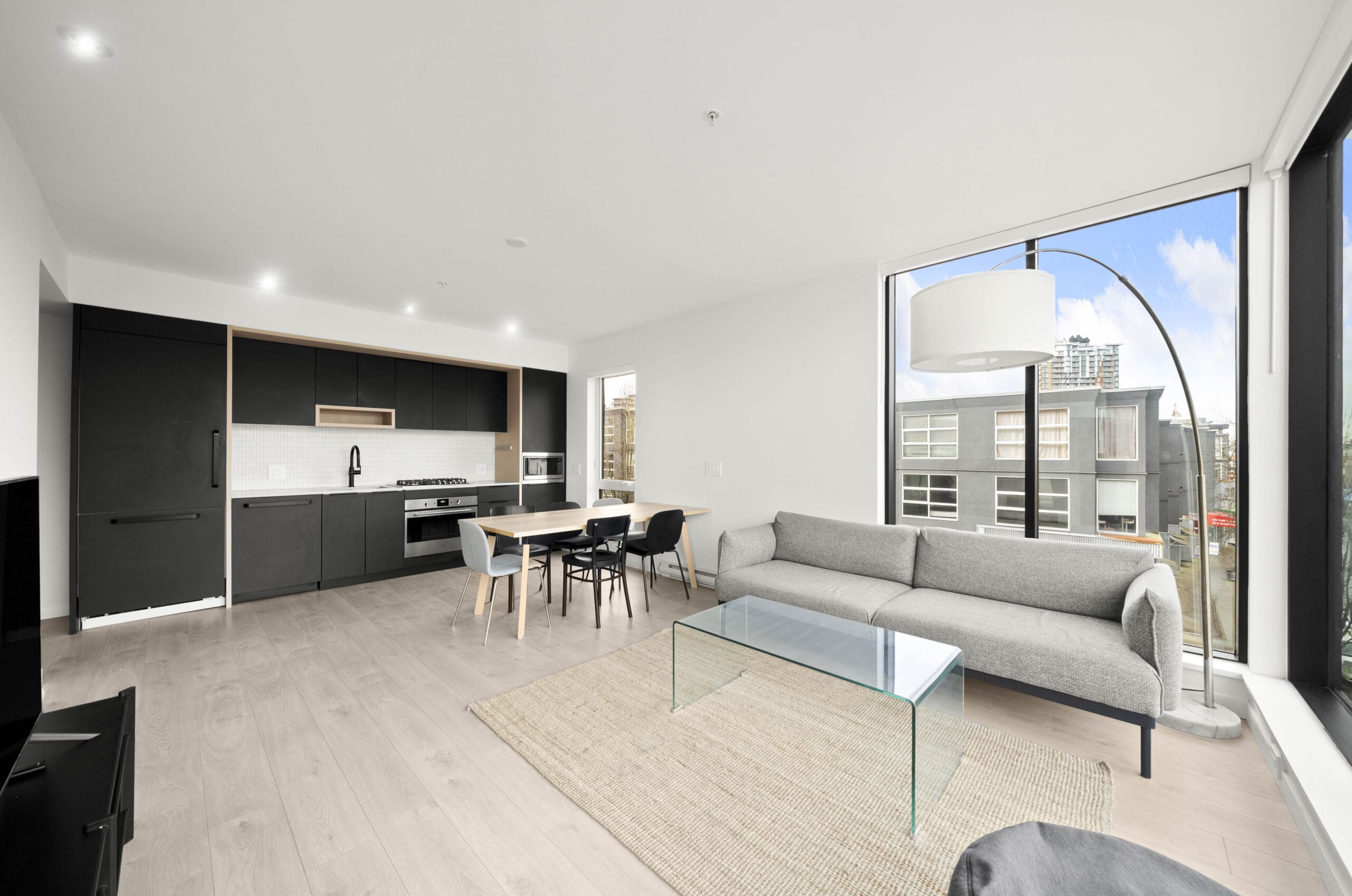Modern open-plan living room and kitchen with black cabinetry, dining table, grey sofa, glass coffee table, large windows, and city views.