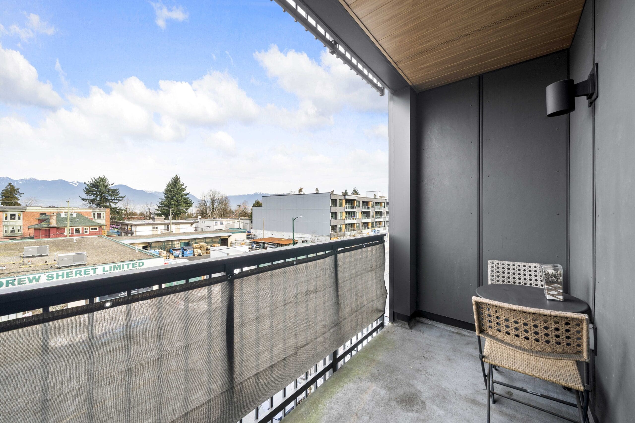 A small balcony with a table and two chairs overlooks a commercial rooftop and distant residential buildings under a partly cloudy sky.