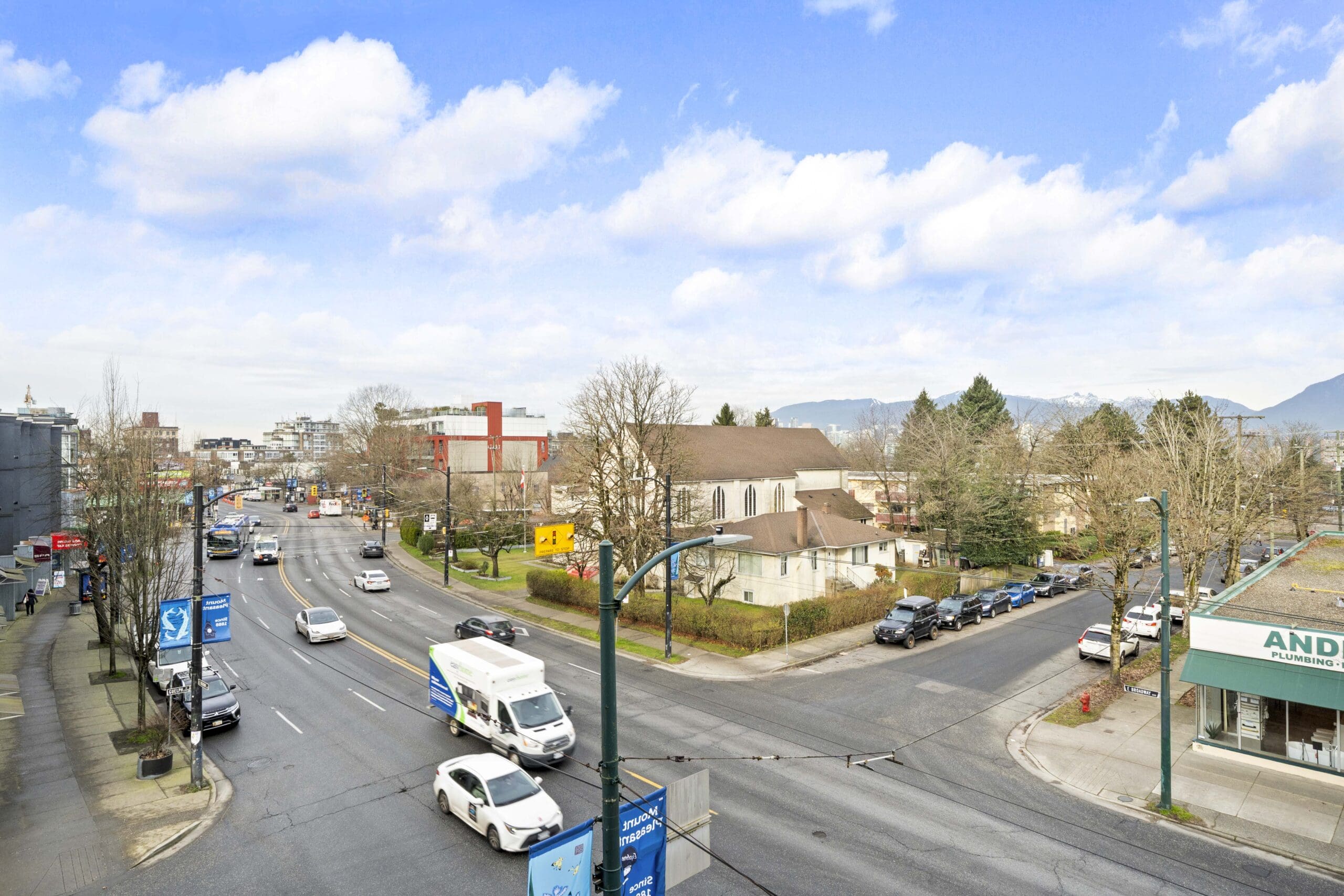 A wide city intersection with cars on the road, surrounding buildings, trees without leaves, and a partly cloudy sky overhead.