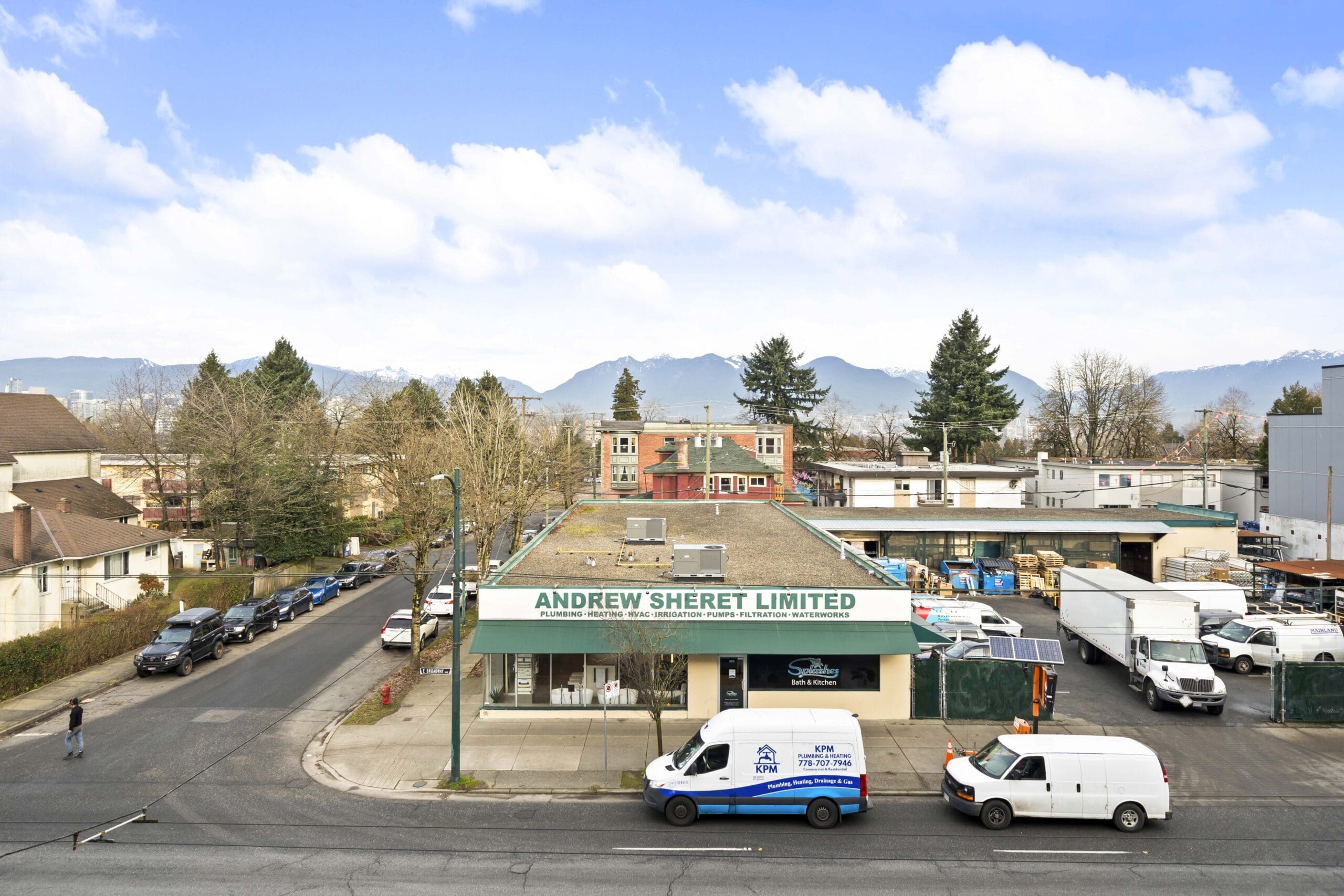 An Andrew Sheret Limited building sits on a street corner with parked vehicles in front; houses, trees, and mountains are visible in the background under a partly cloudy sky.