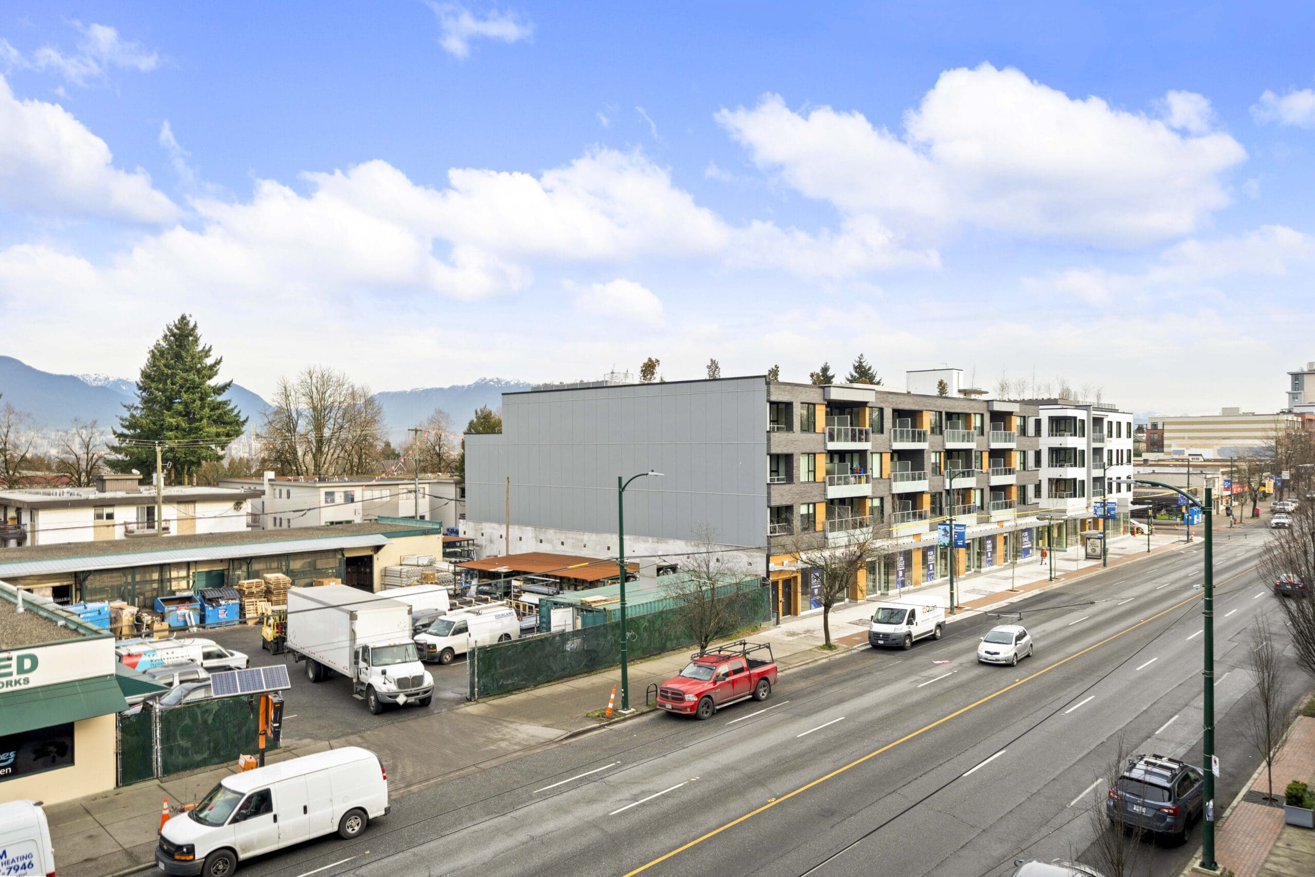 A view of a busy urban street with vehicles parked and driving, commercial buildings, an apartment complex, and some trees under a partly cloudy sky.