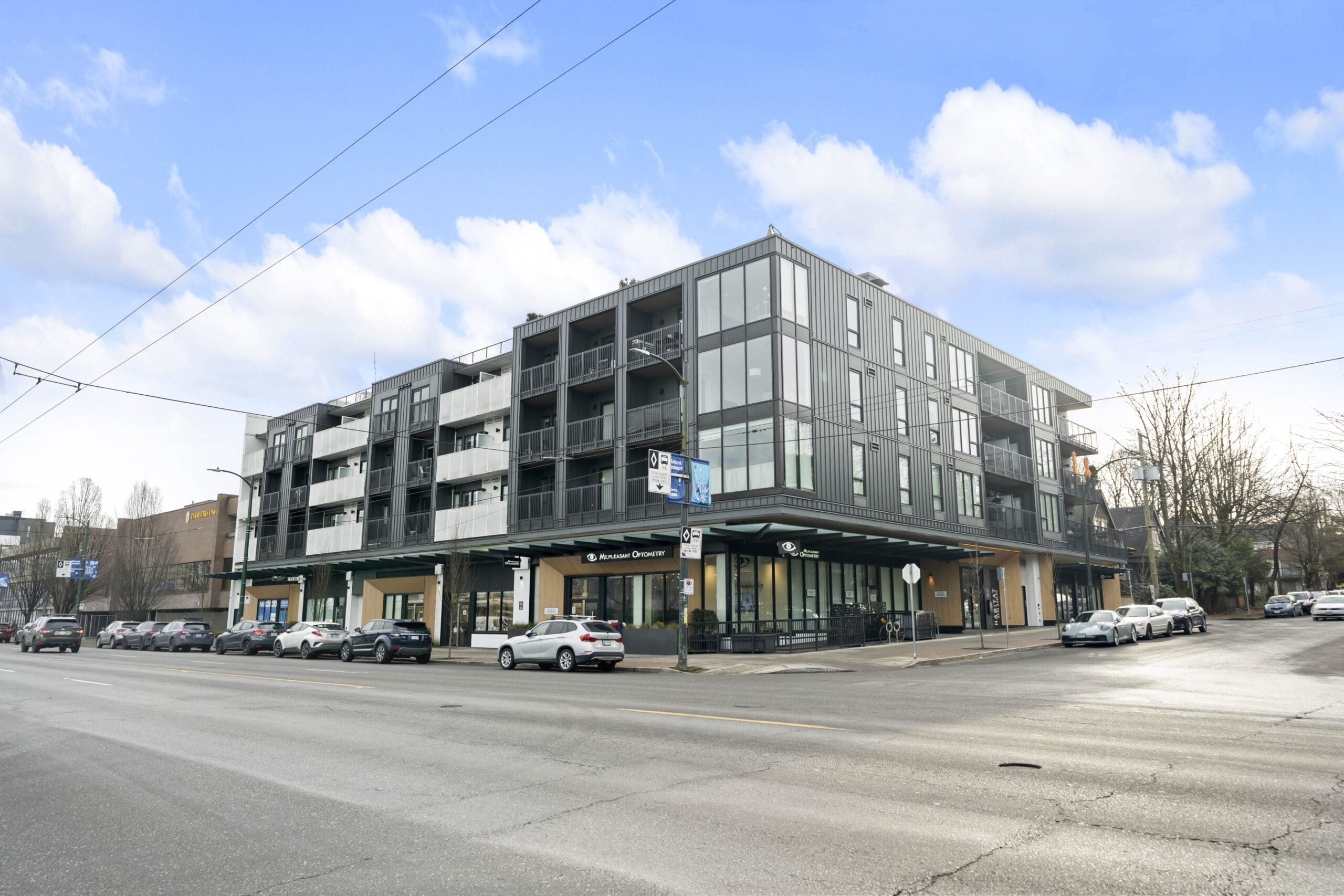 Modern four-story apartment building with balconies and ground-floor retail spaces on a city street corner, with parked cars along the road.