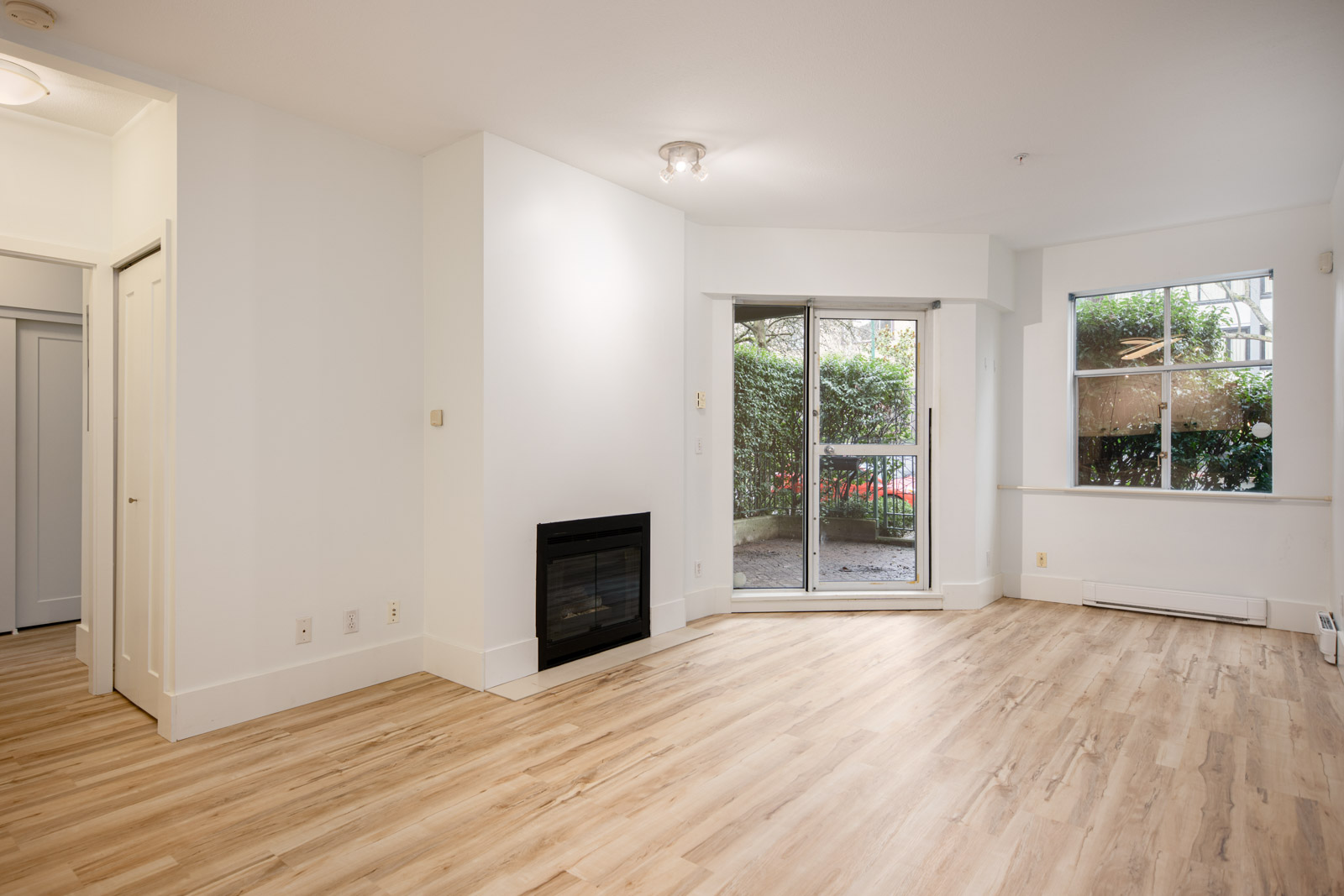 Unfurnished living room with light wood flooring, white walls, a fireplace, sliding glass doors to a patio, and a large window facing greenery.