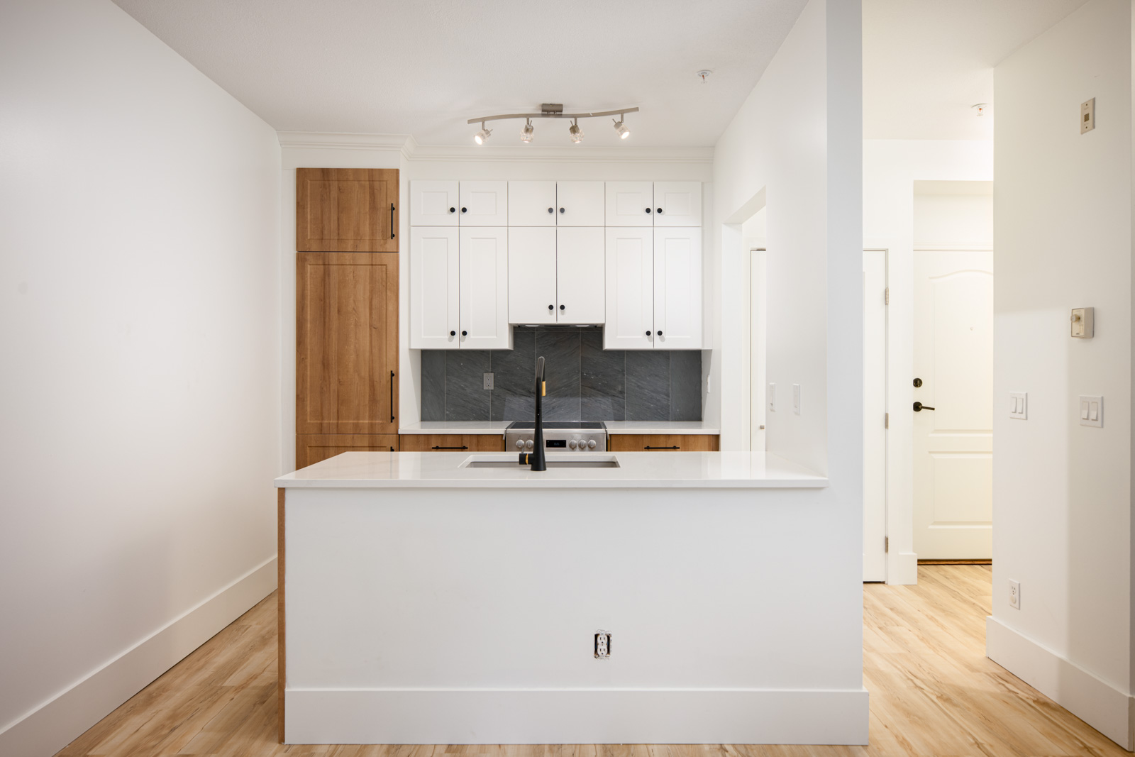 Modern kitchen with white cabinets, wood accents, a central island with sink, black faucet, and overhead track lighting on a light hardwood floor.