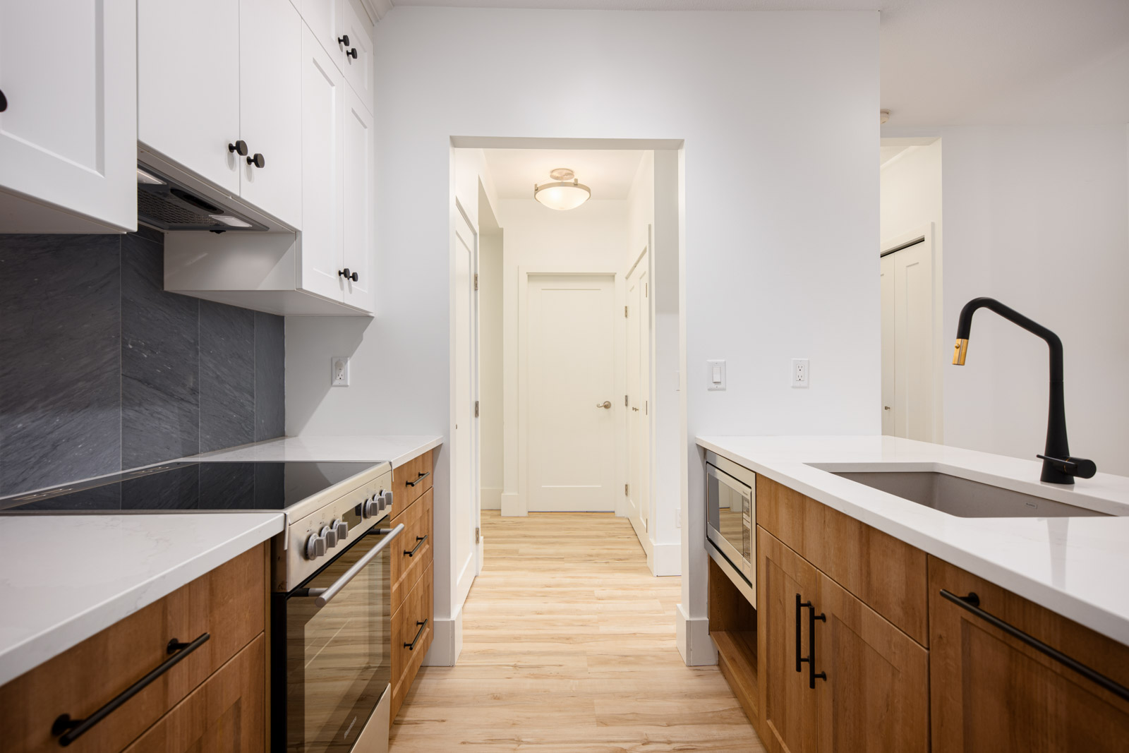 Modern kitchen with wooden lower cabinets, white upper cabinets, stainless steel appliances, and a black faucet, opening to a hallway with light wood flooring and white walls.