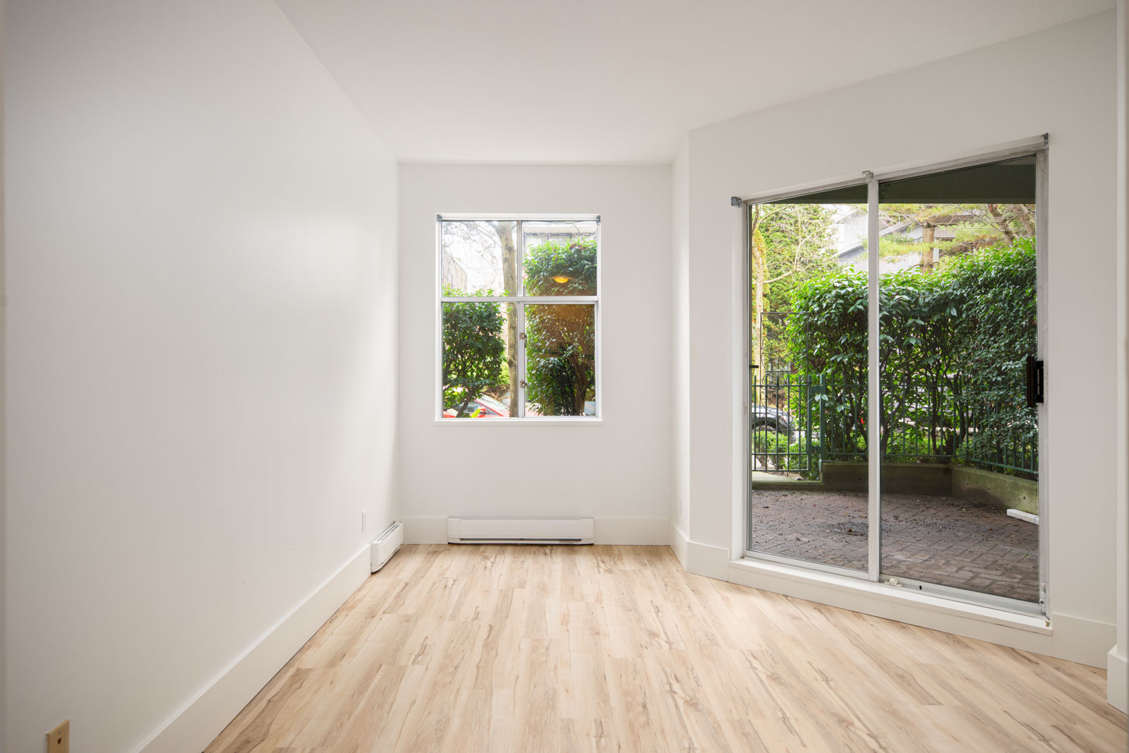Empty, unfurnished room with light wood flooring, white walls, a window, and a sliding glass door leading to a patio with greenery outside.