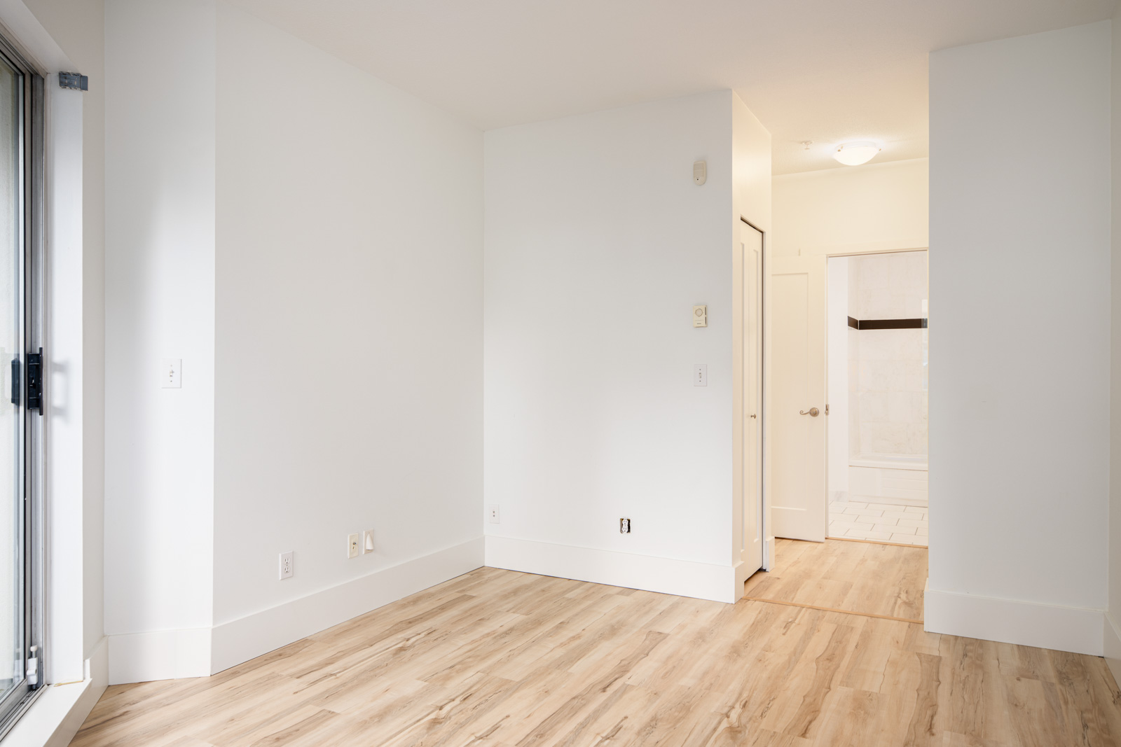 An empty room with light-colored wood flooring, white walls, a small hallway leading to a bathroom, and a large window on the left side.