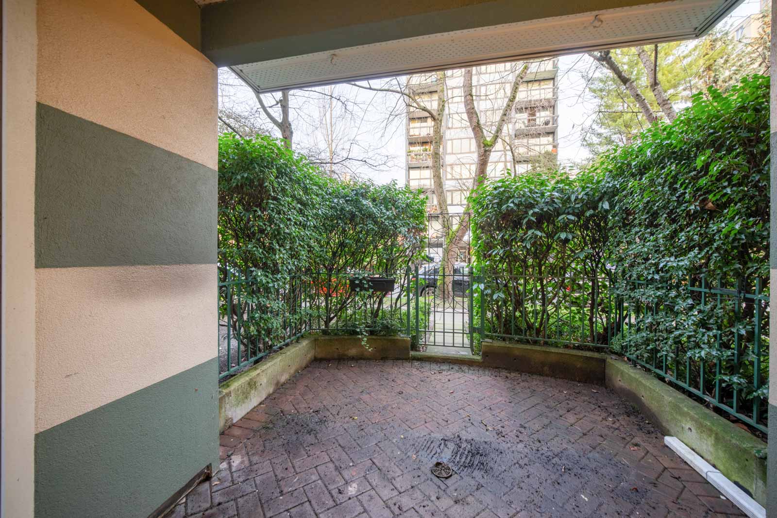 Small brick patio with metal gate, surrounded by green shrubs and a fence, adjacent to a multi-story apartment building.