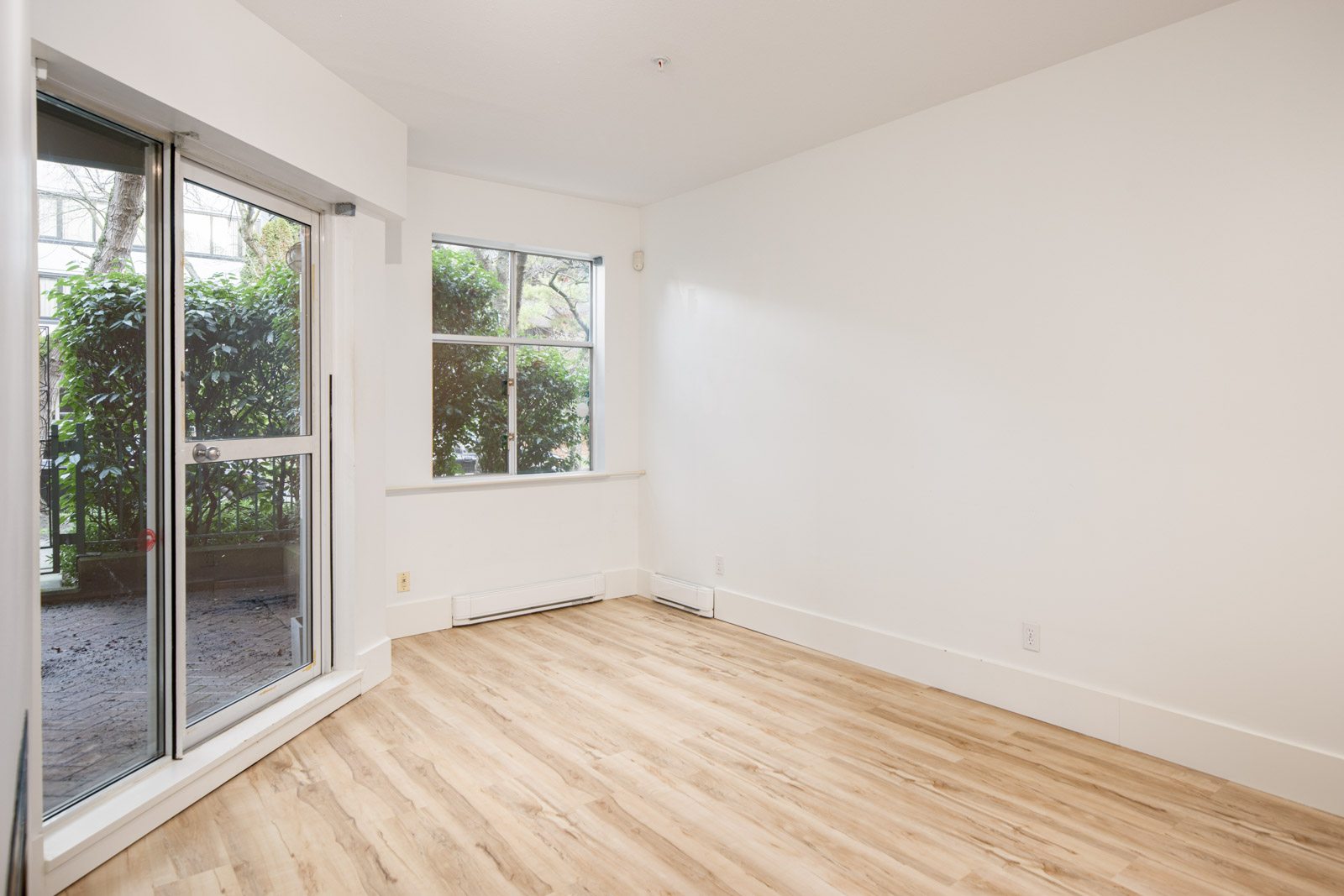 Empty room with light wood flooring, white walls, a large window, a glass door, and view of greenery outside.