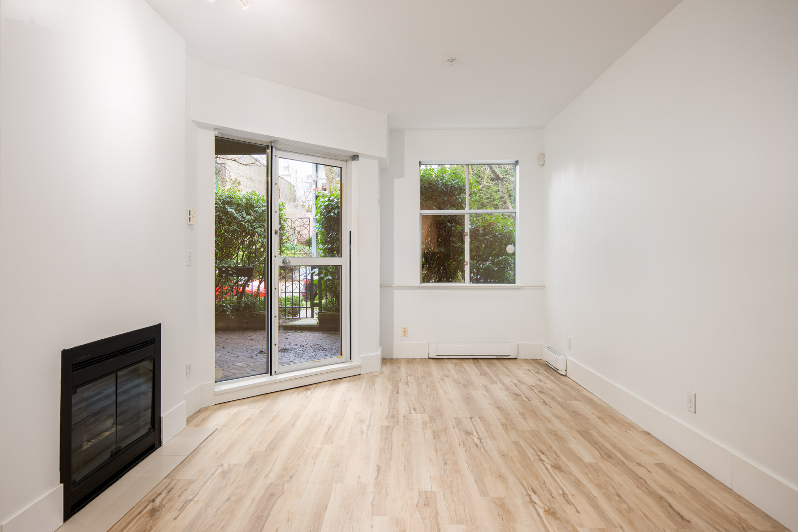 Unfurnished living room with light wood flooring, a fireplace on the left, sliding glass doors, and a large window overlooking a patio and greenery.