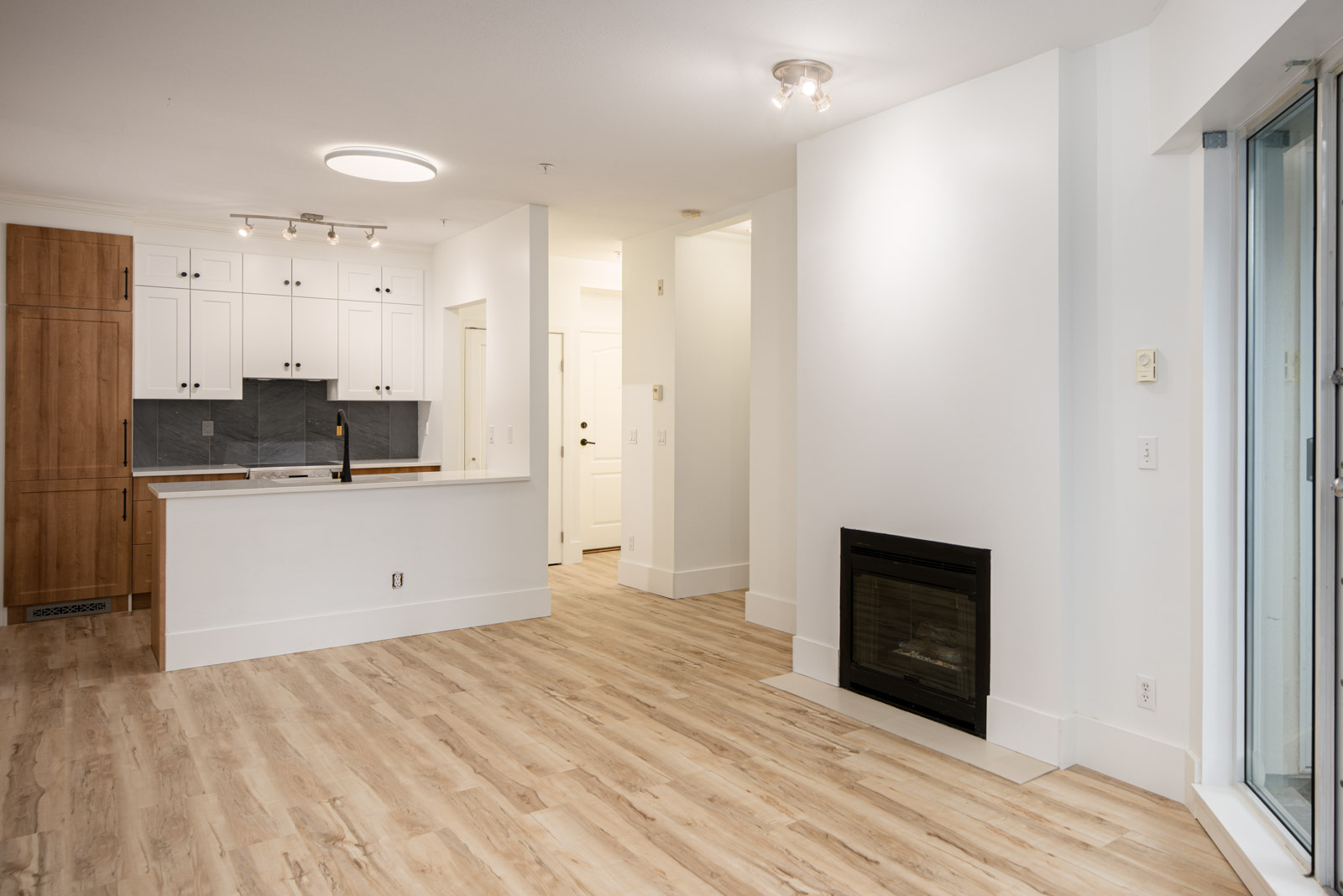 Modern apartment interior with light wood flooring, white walls, open kitchen with white cabinets, black sink, built-in fireplace, and sliding glass door leading outside.
