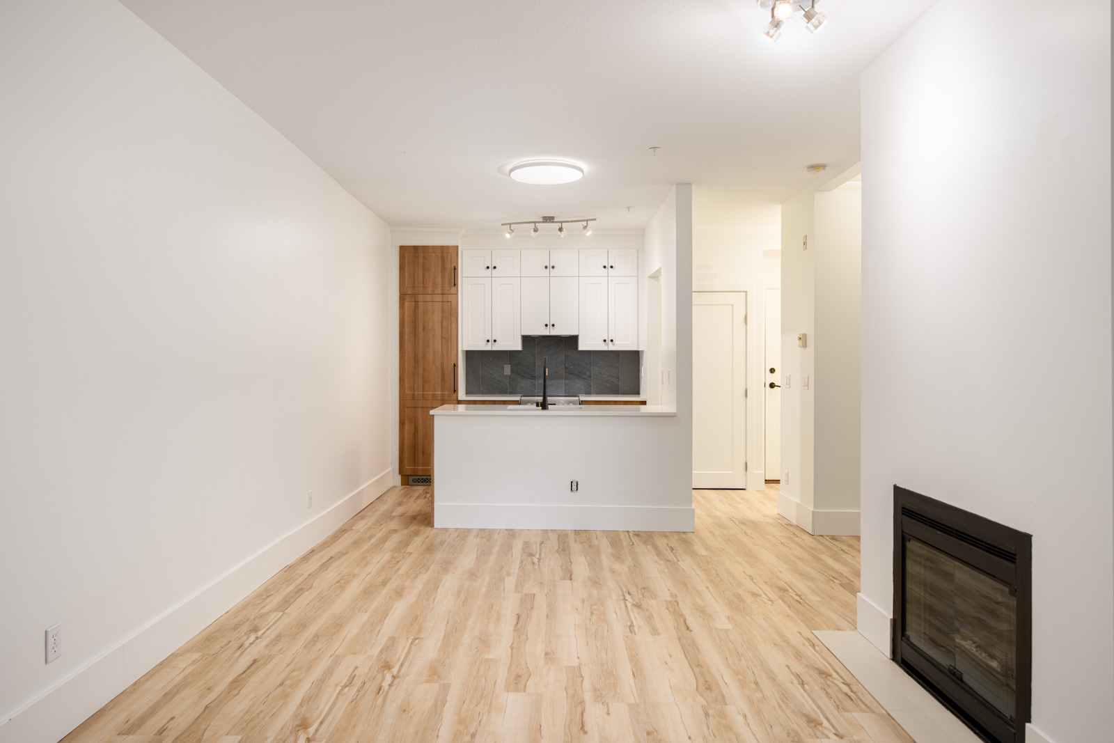 Unfurnished open-plan living area with light wood flooring, white walls, a modern fireplace, and a kitchen featuring white cabinets and a wood accent pantry.