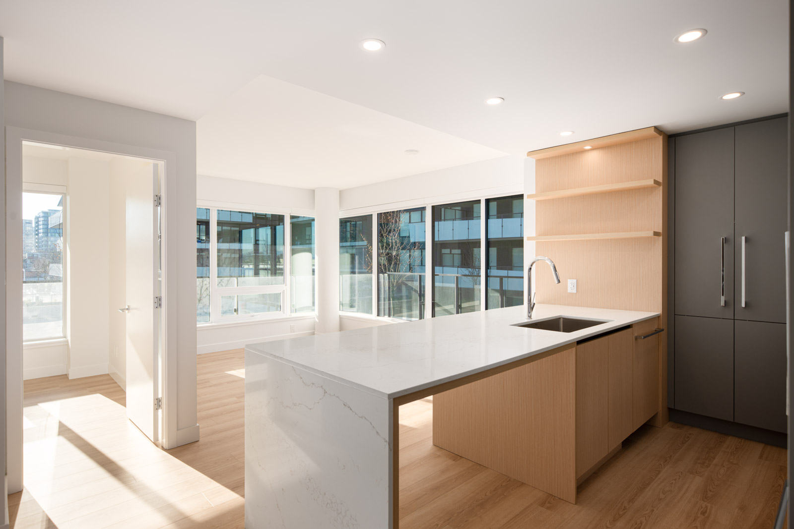 Modern kitchen with a large marble island, wood cabinetry, built-in shelves, and tall windows letting in natural light; adjacent room visible through an open door.