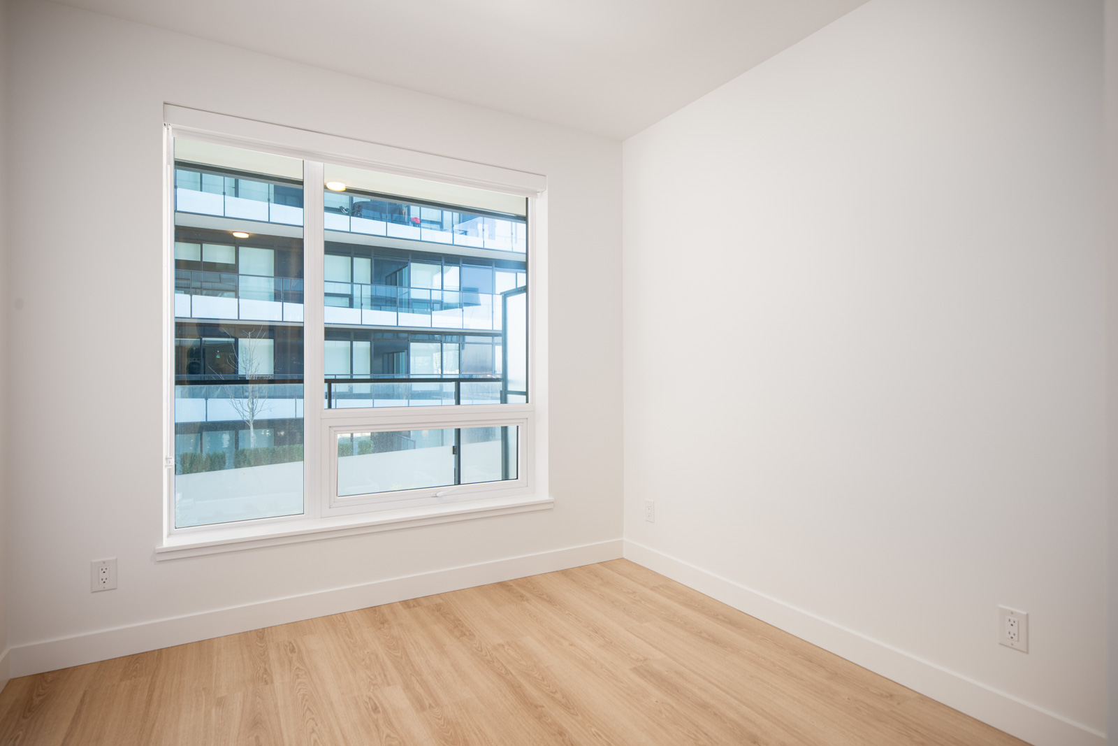 Empty room with white walls, large window, and light wood flooring; exterior of a modern building with balconies visible through the window.