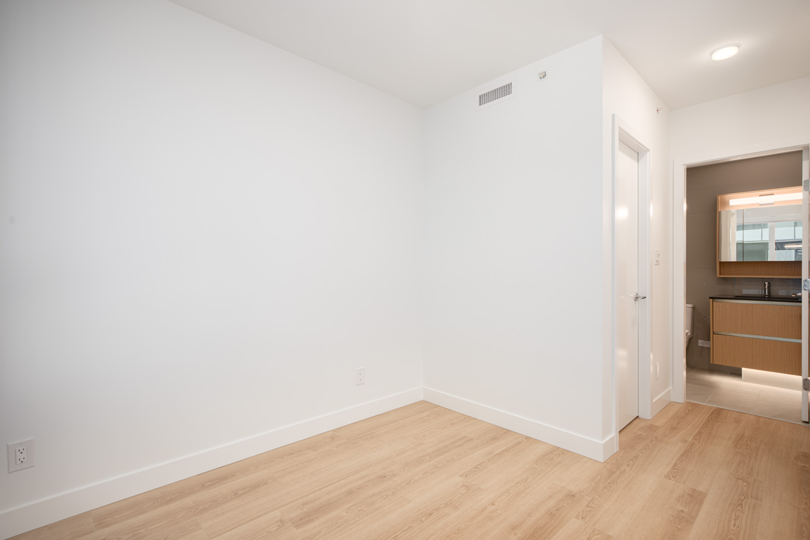 Empty room with white walls, light wood flooring, and a door leading to a bathroom with a lit mirror and vanity visible.