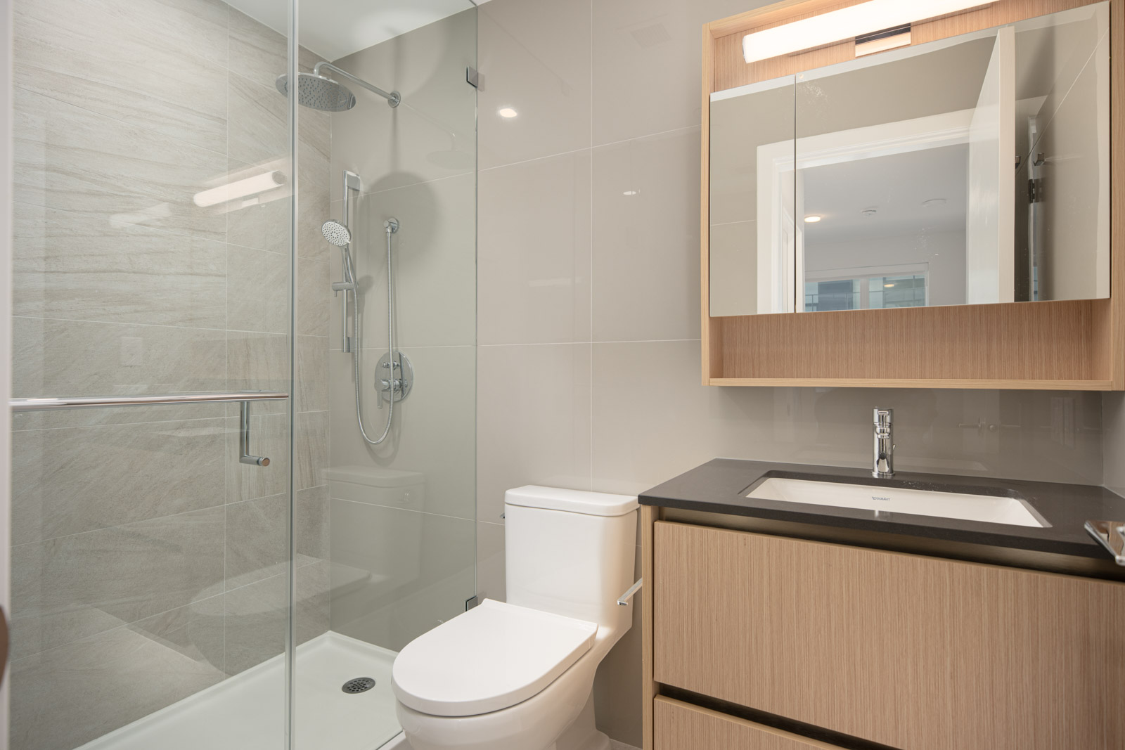 Modern bathroom with a glass-enclosed shower, white toilet, and wood vanity with a rectangular sink and mirror cabinet above.