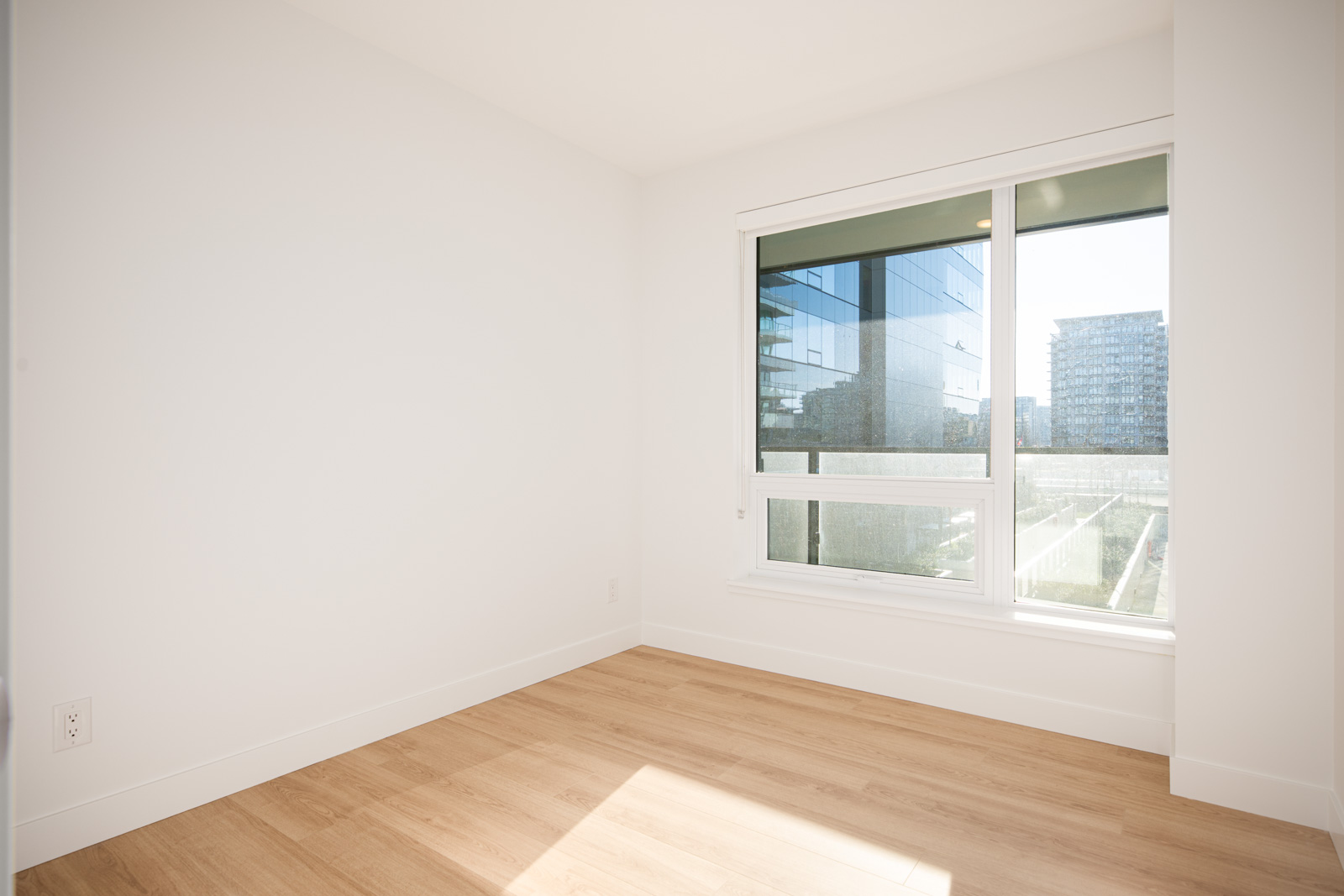 Empty, unfurnished room with white walls, light wood flooring, and a large window overlooking modern buildings and an outdoor area.