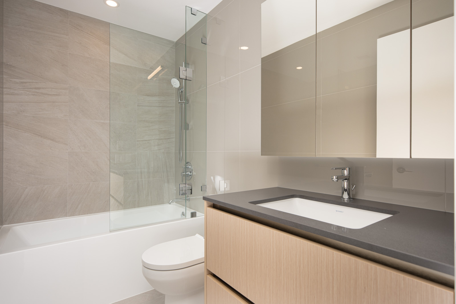 Modern bathroom with a glass-enclosed shower, built-in bathtub, wall-mounted showerhead, light wood vanity, black countertop, and large mirror cabinets.