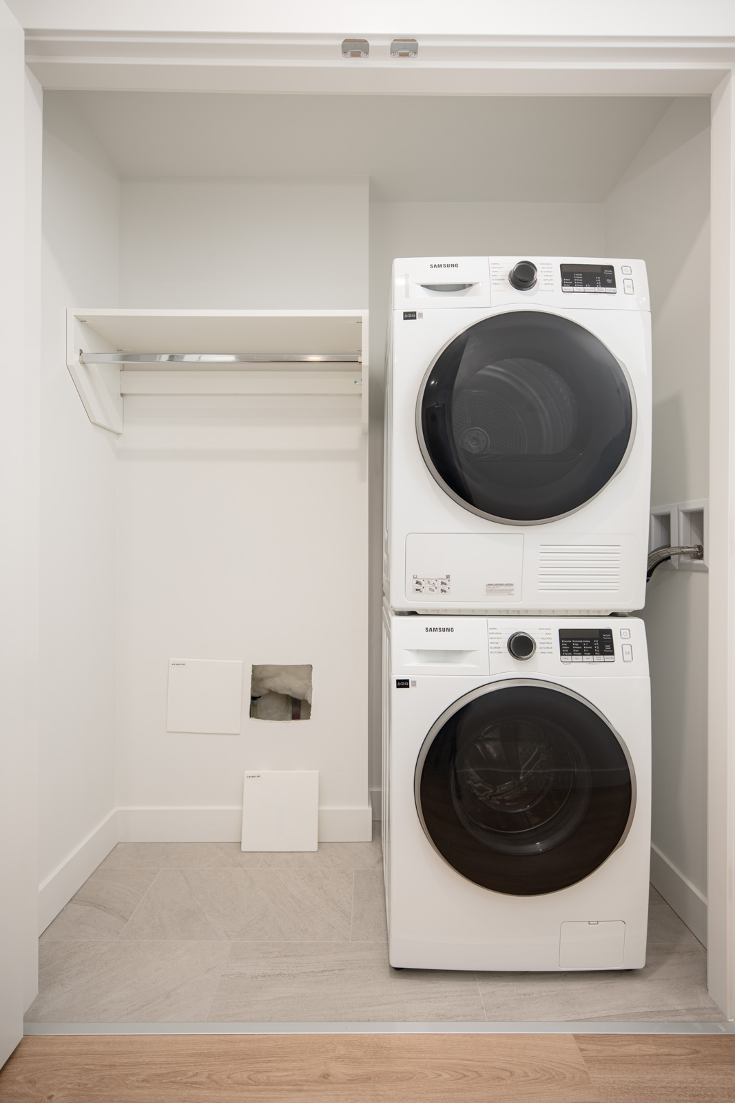 A white stacked Samsung washer and dryer set is positioned in a small laundry nook with a shelf, hanging rod, and paint color samples on the floor.