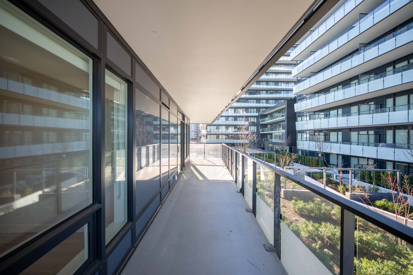 View of a modern apartment building hallway with glass railings, overlooking a landscaped courtyard and adjacent buildings.