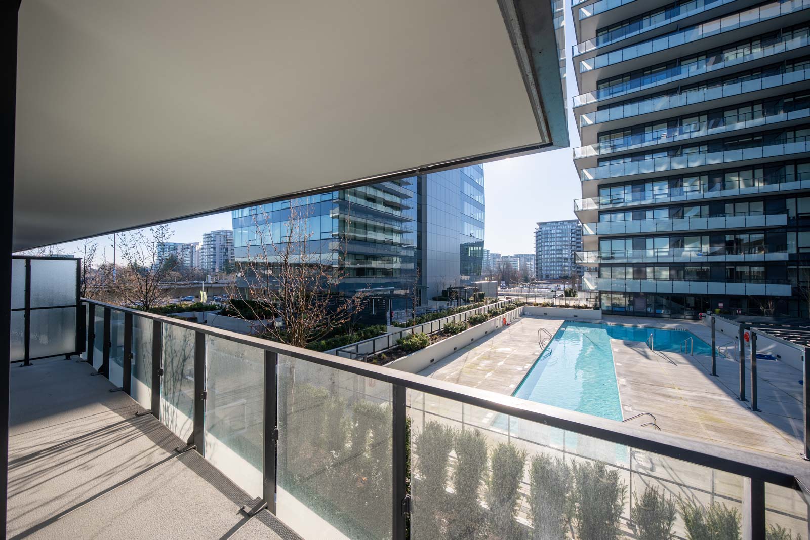 View from a balcony overlooking a swimming pool and modern glass apartment buildings on a sunny day.