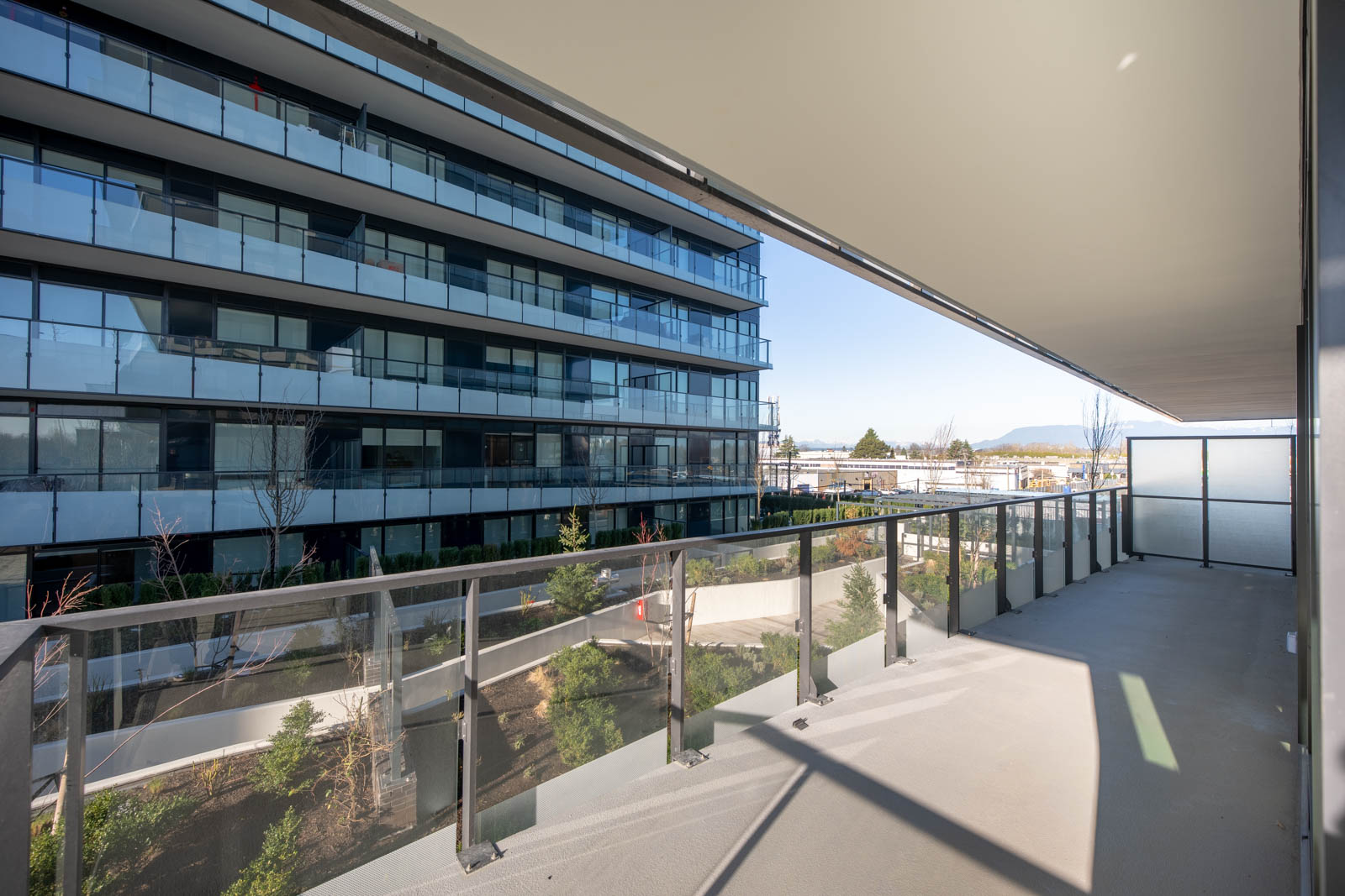 A spacious balcony with glass railing overlooks a landscaped courtyard and a modern multi-story apartment building on a clear day.