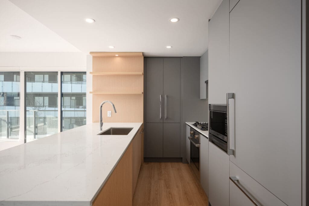 Modern kitchen with grey cabinets, built-in oven, gas stove, wood floors, and a large white island countertop with a sink and faucet. Large windows provide natural light.