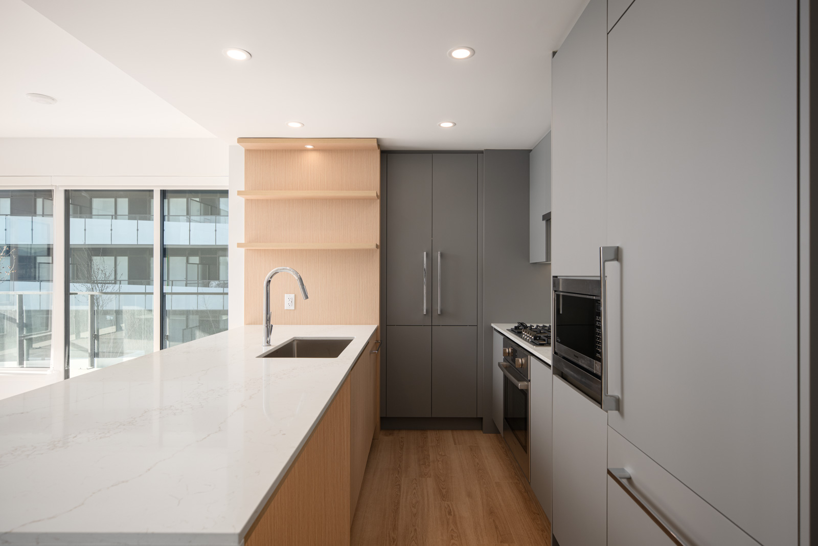 Modern kitchen with grey cabinets, built-in oven, gas stove, wood floors, and a large white island countertop with a sink and faucet. Large windows provide natural light.