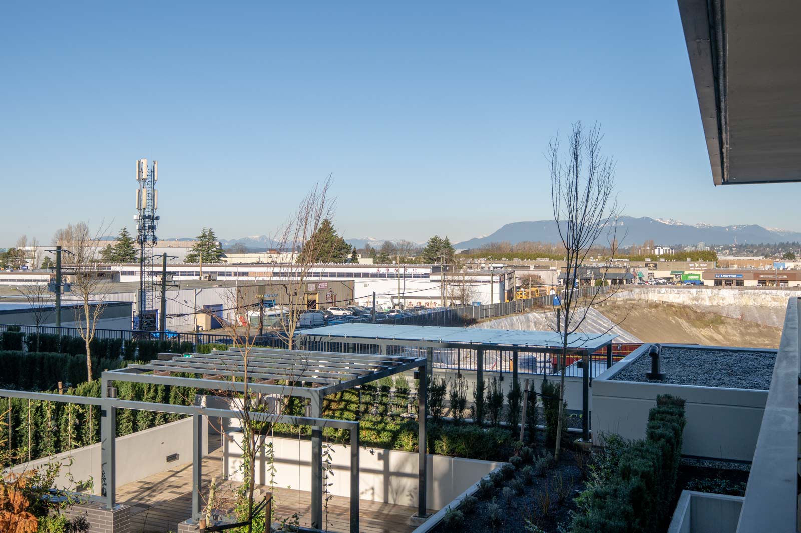 View of an industrial area with warehouses, a cell tower, and mountains in the distance, seen from a building with a landscaped garden terrace in the foreground.