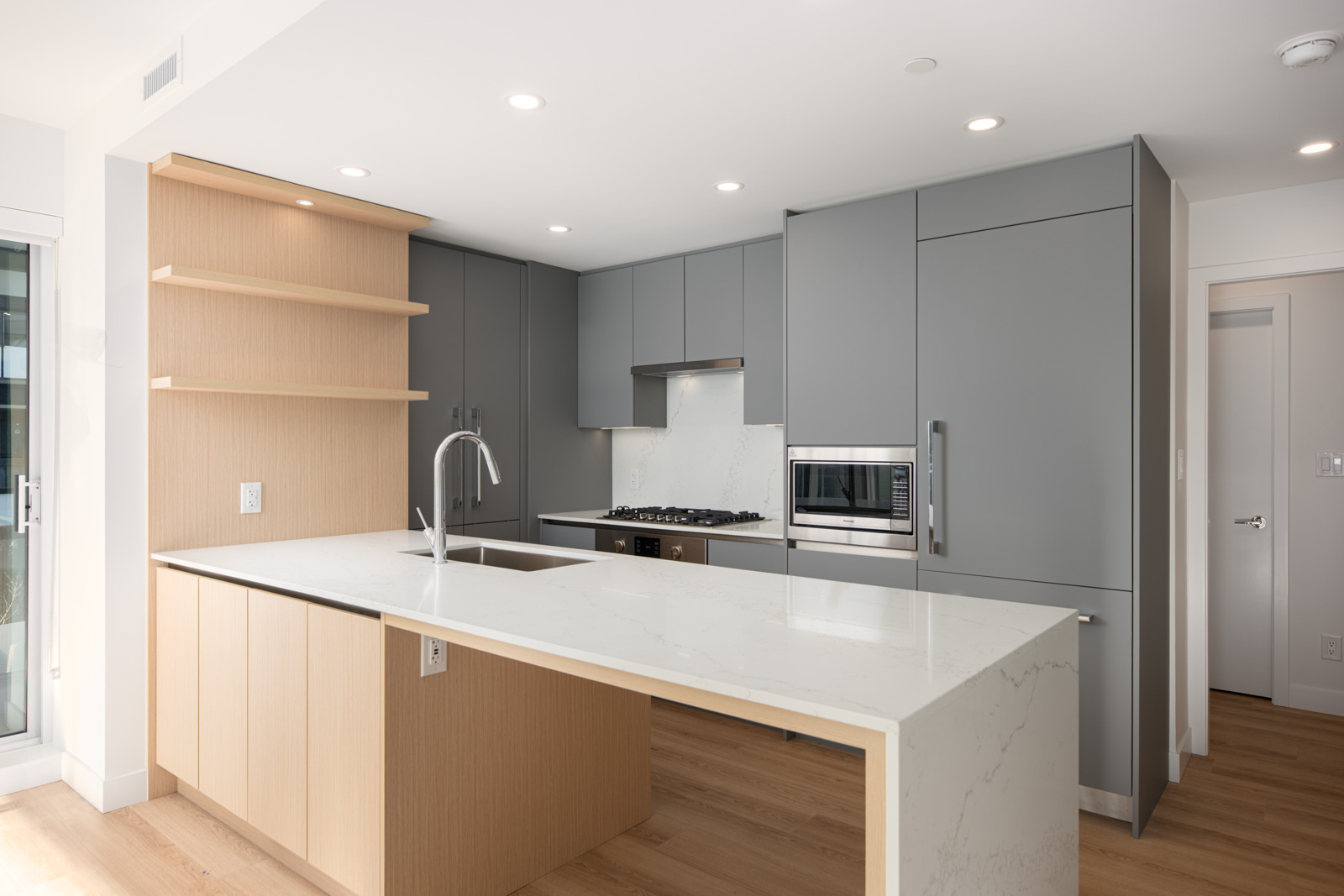 Modern kitchen with light wood and gray cabinets, white marble countertops, built-in appliances, and open shelving, featuring a large island and recessed lighting.