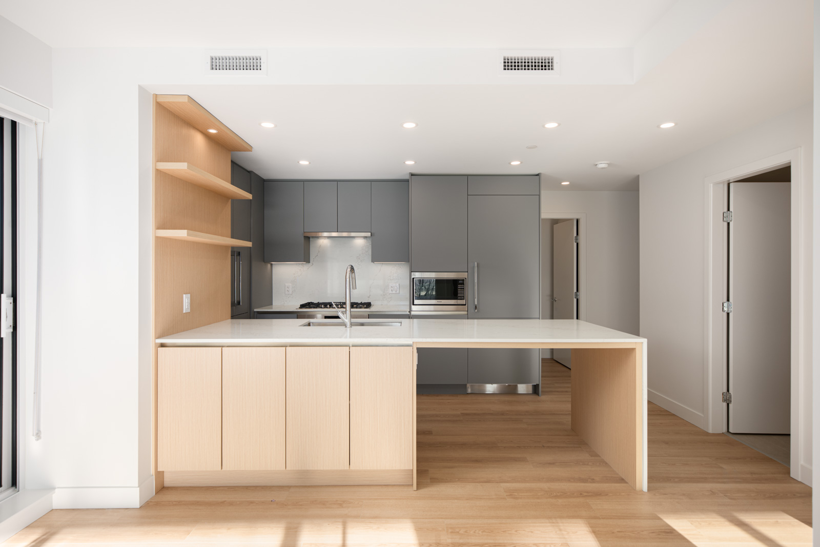 Modern kitchen with gray cabinets, light wood accents, an island with a built-in sink, open shelving, and stainless steel appliances on light wood flooring.