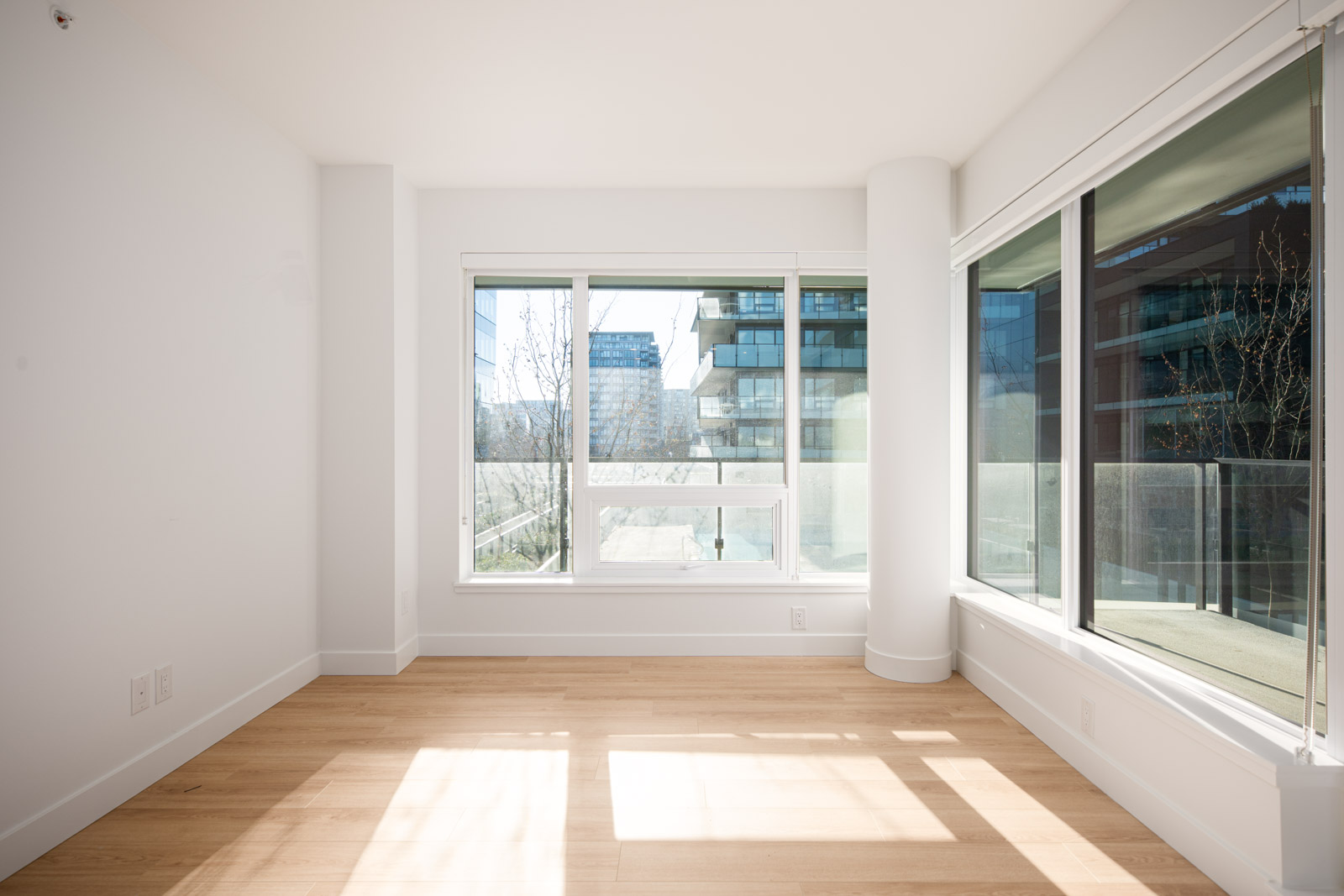 Bright, empty apartment room with large windows and light wood flooring, overlooking modern buildings and a balcony outside.