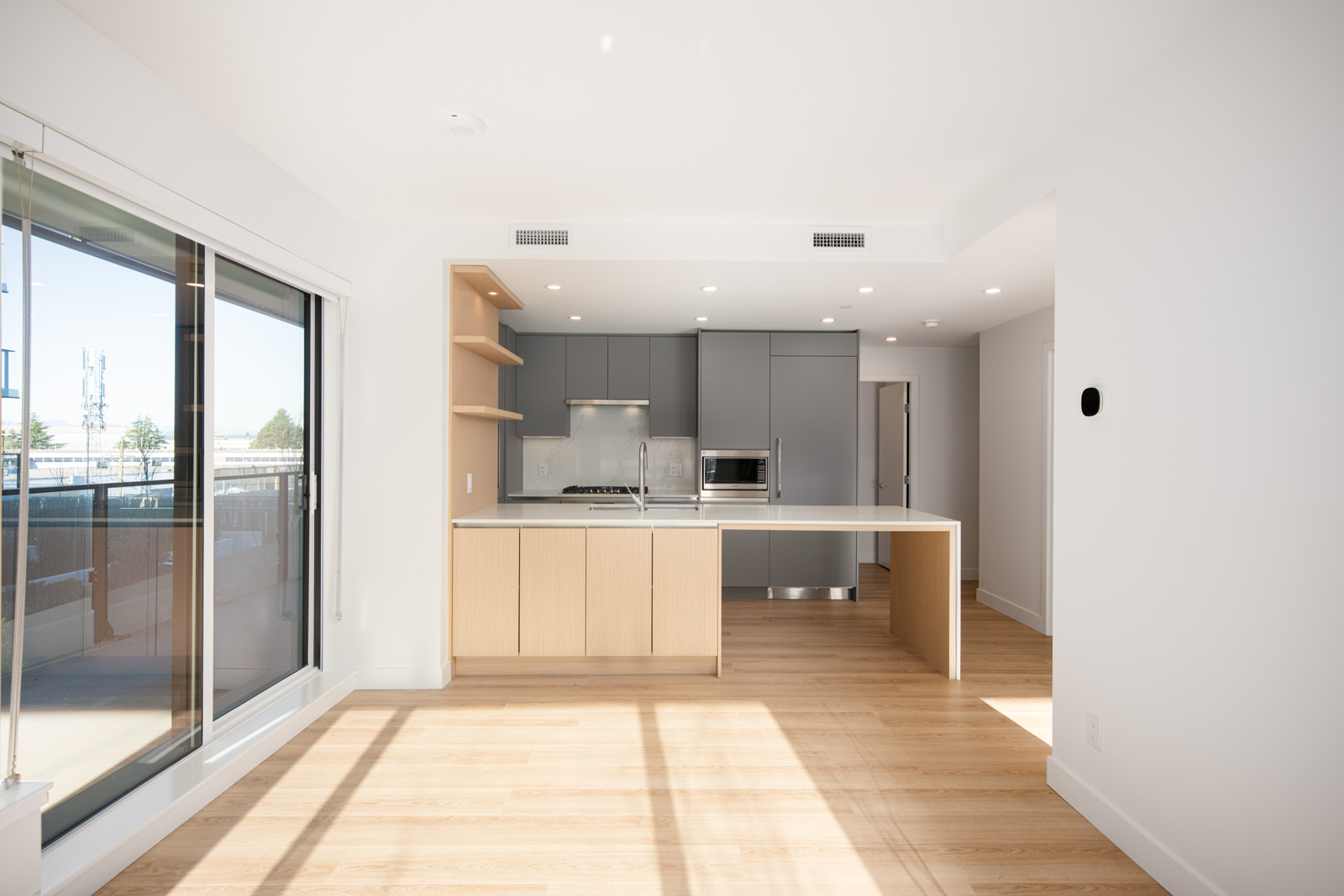 Modern, unfurnished kitchen with gray cabinets, stainless steel appliances, light wood flooring, an island, and large windows allowing natural light into the space.