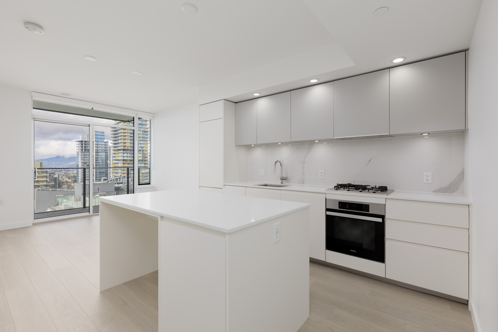 Modern, minimalist kitchen with white cabinetry and countertops, built-in oven, gas stove, and an island. Floor-to-ceiling windows reveal a balcony and city buildings outside.