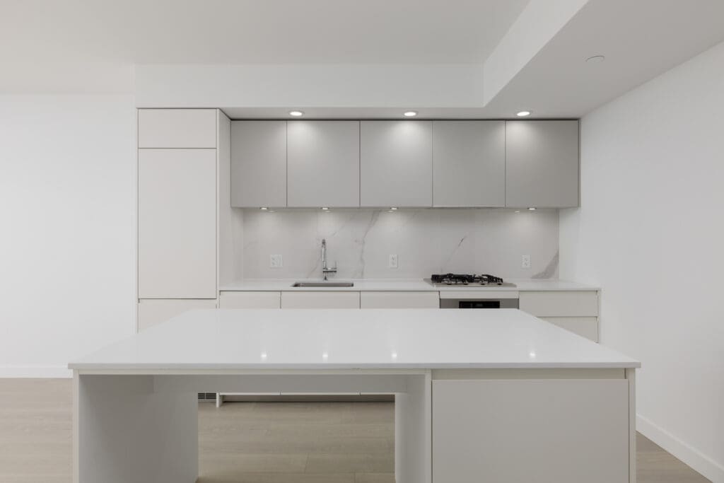 Minimalist modern kitchen with light grey cabinets, white countertops, built-in sink, stovetop, and under-cabinet lighting. Large island in the foreground.