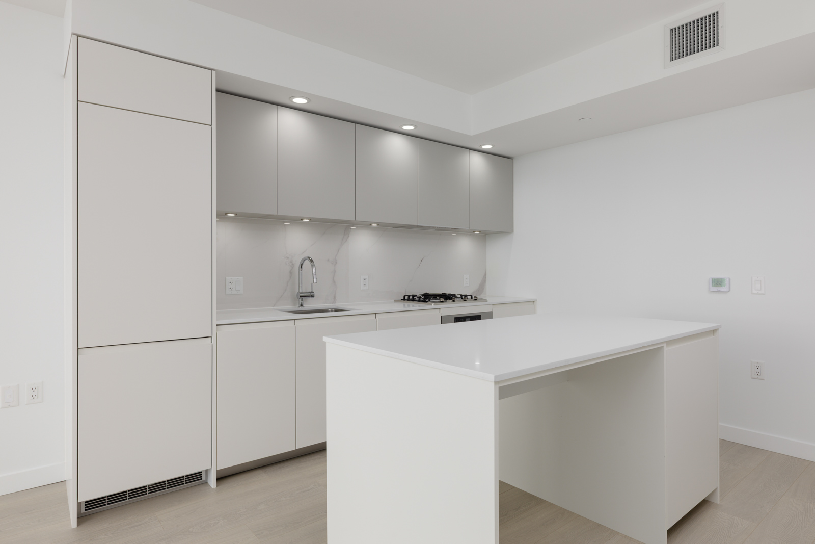 Minimalist modern kitchen with white cabinetry, built-in appliances, under-cabinet lighting, marble backsplash, and a central island on light wood flooring.