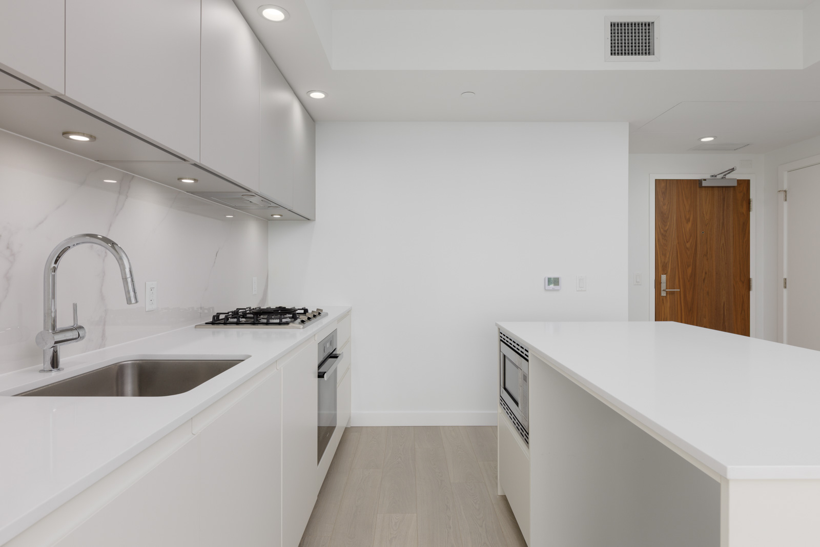 Modern kitchen with white cabinets, a built-in oven, gas cooktop, stainless steel sink, and a large white island; wooden door visible in the background.