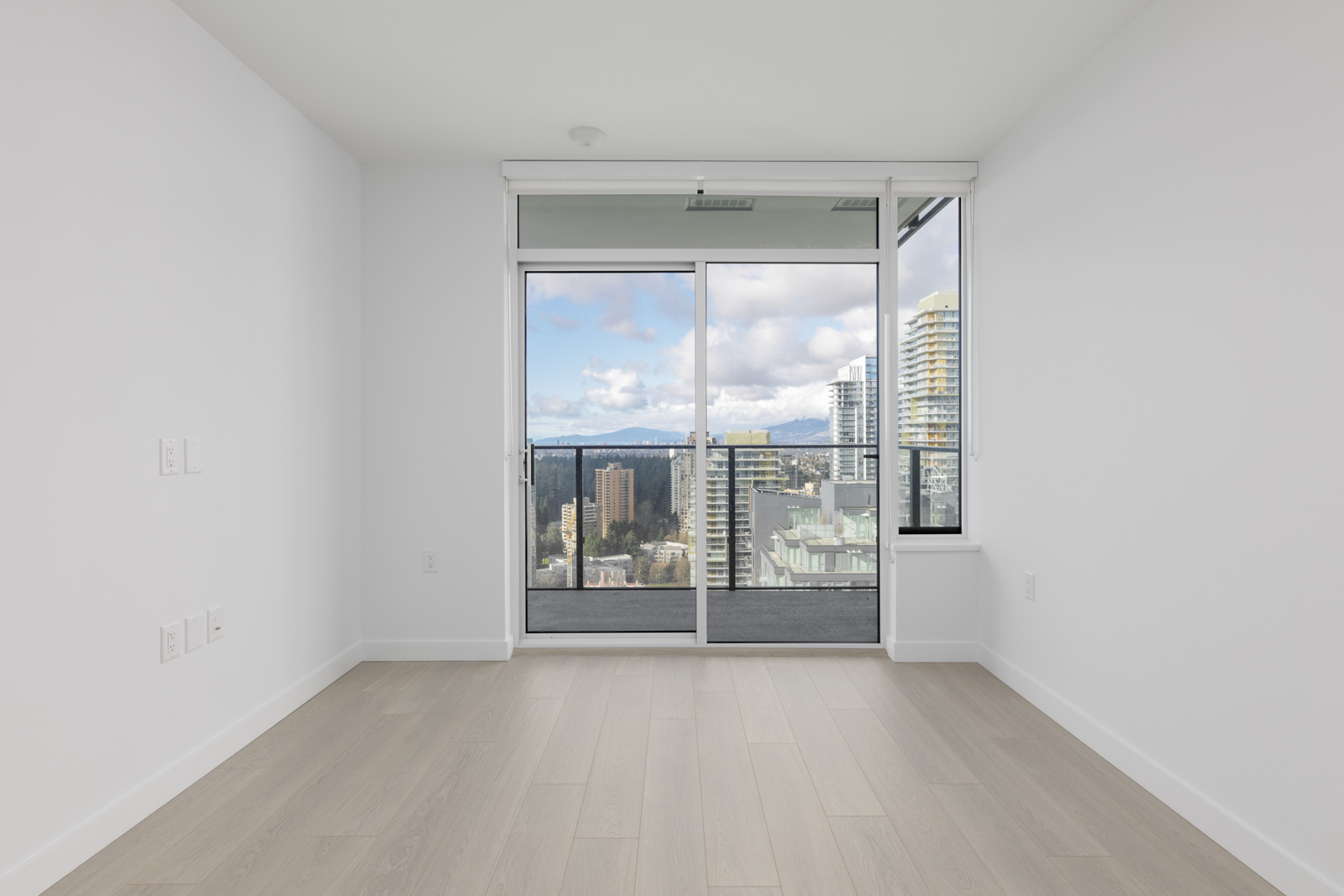Unfurnished room with light wood flooring, white walls, and a glass door leading to a balcony overlooking a cityscape with high-rise buildings and mountains in the background.